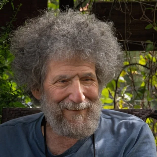Older man with gray, curly hair and beard smiling outdoors surrounded by greenery.