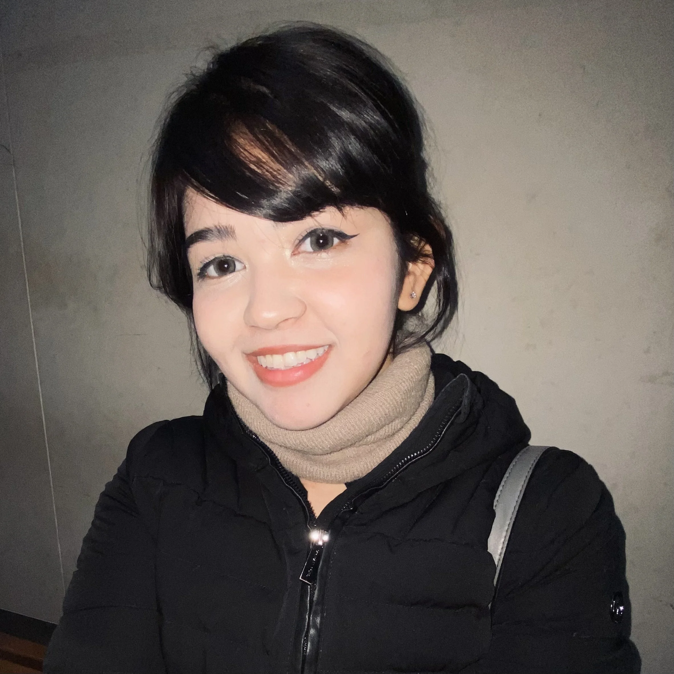 A young woman with short dark hair, smiling, wearing a beige turtleneck and black jacket, with a gray strap on her shoulder, standing against a plain wall.