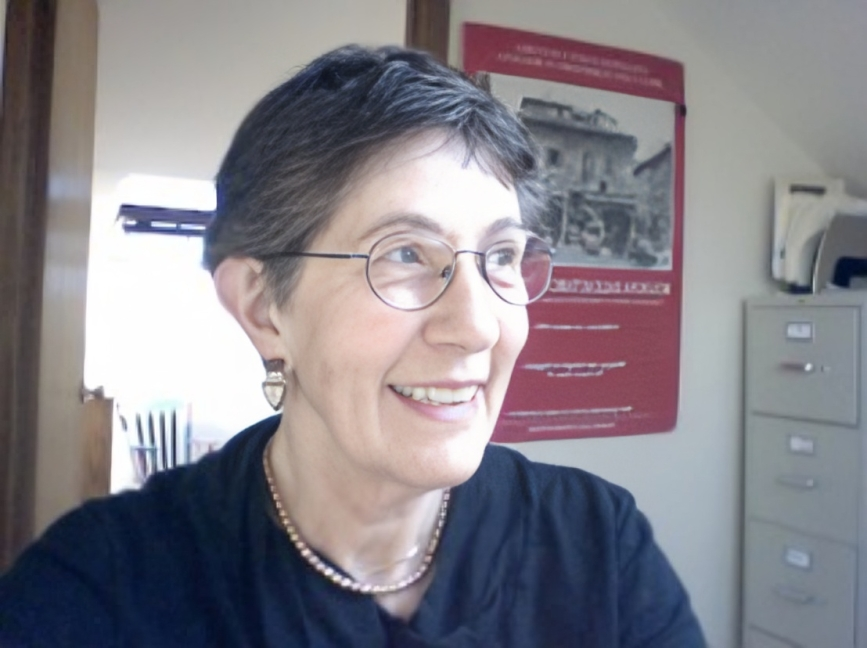 A woman with short brown hair and glasses smiling, wearing a black top and a pearl necklace, in an indoor office setting.