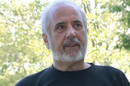 A man with gray hair and a beard outdoors with trees and sunlight in the background.