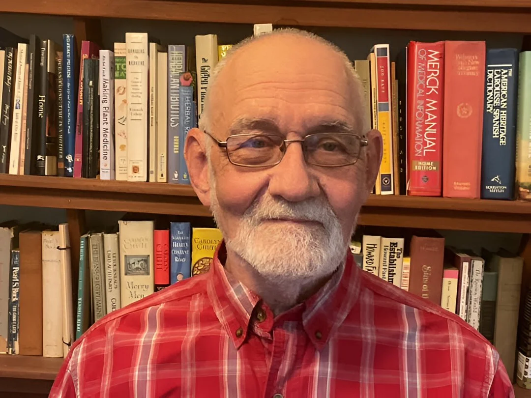 An elderly man with glasses and a white beard and mustache wearing a red plaid shirt stands in front of bookshelves filled with a variety of books.