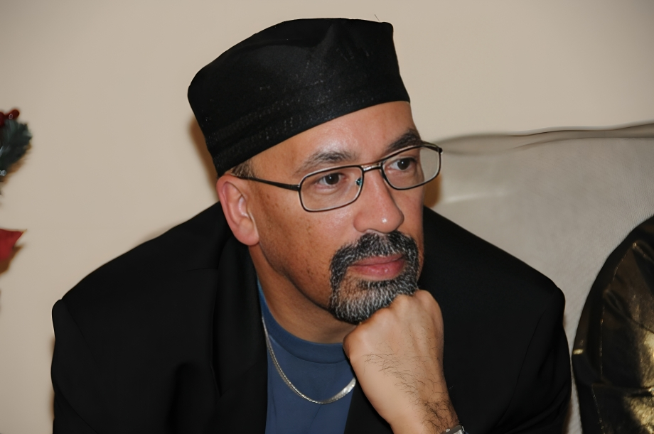 A man wearing glasses, a black cap, and a black blazer, sitting with his hand under his chin, looking thoughtful.