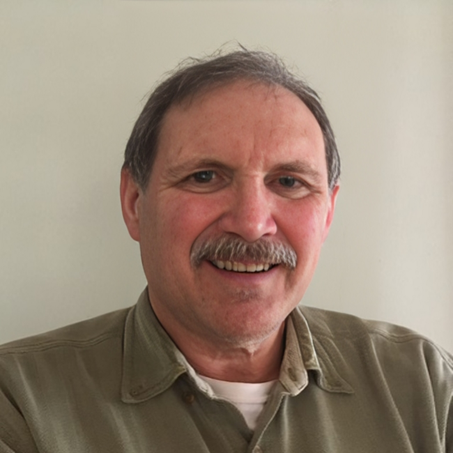 A middle-aged man with gray hair and a gray mustache smiling, wearing a tan shirt, against a plain light-colored wall.