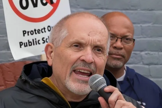 Older man speaking into a microphone with a serious expression, standing outdoors with another man in the background near a protest sign.