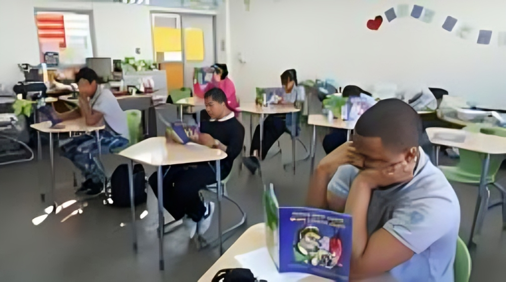Students in a classroom, sitting at desks reading books with their faces buried in them, spaced apart.