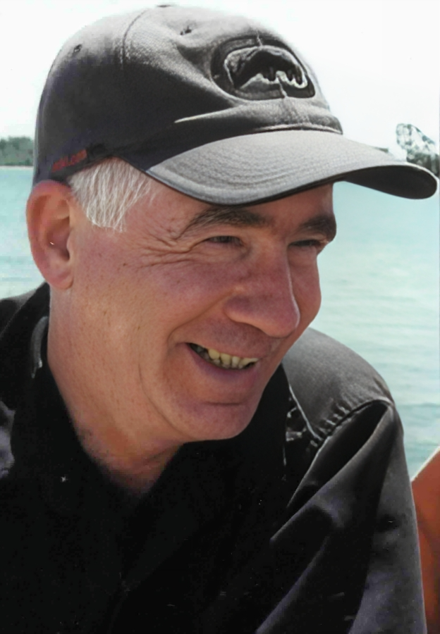 A smiling man wearing a gray baseball cap and black jacket, outdoors near water.