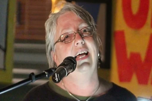 A person with short gray hair and glasses speaking or singing into a microphone, with a blurred background that includes a yellow and red sign.