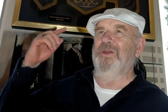 Older man with a white beard and mustache wearing a white chef's hat and dark jacket, pointing to his head with his index finger, standing indoors.