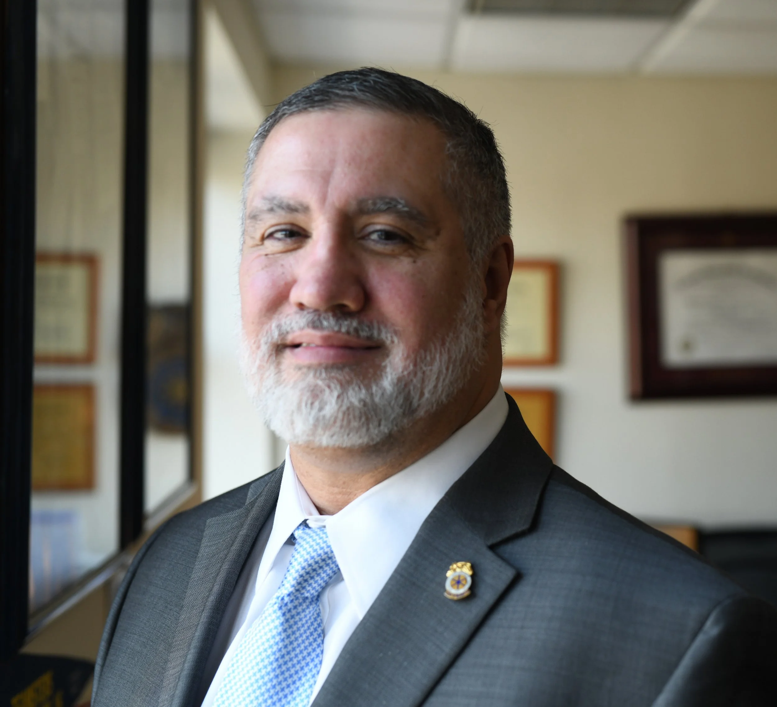 A middle-aged man with gray hair and a beard, dressed in a dark suit with a light blue tie, standing indoors near a window with framed certificates or awards in the background.