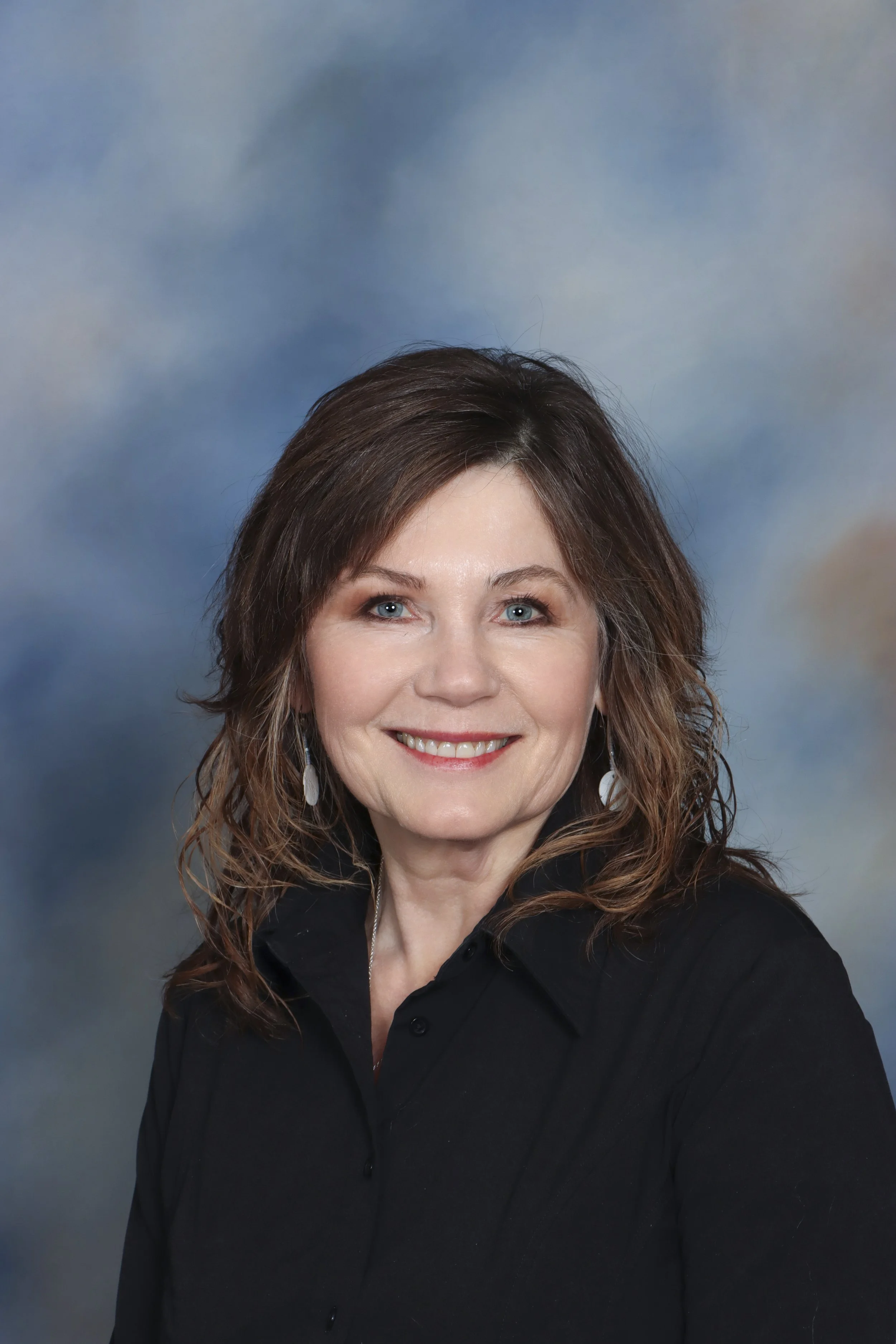A portrait of a woman with shoulder-length brown hair wearing a brown button-up shirt and a blue beaded necklace, smiling against a gray background.