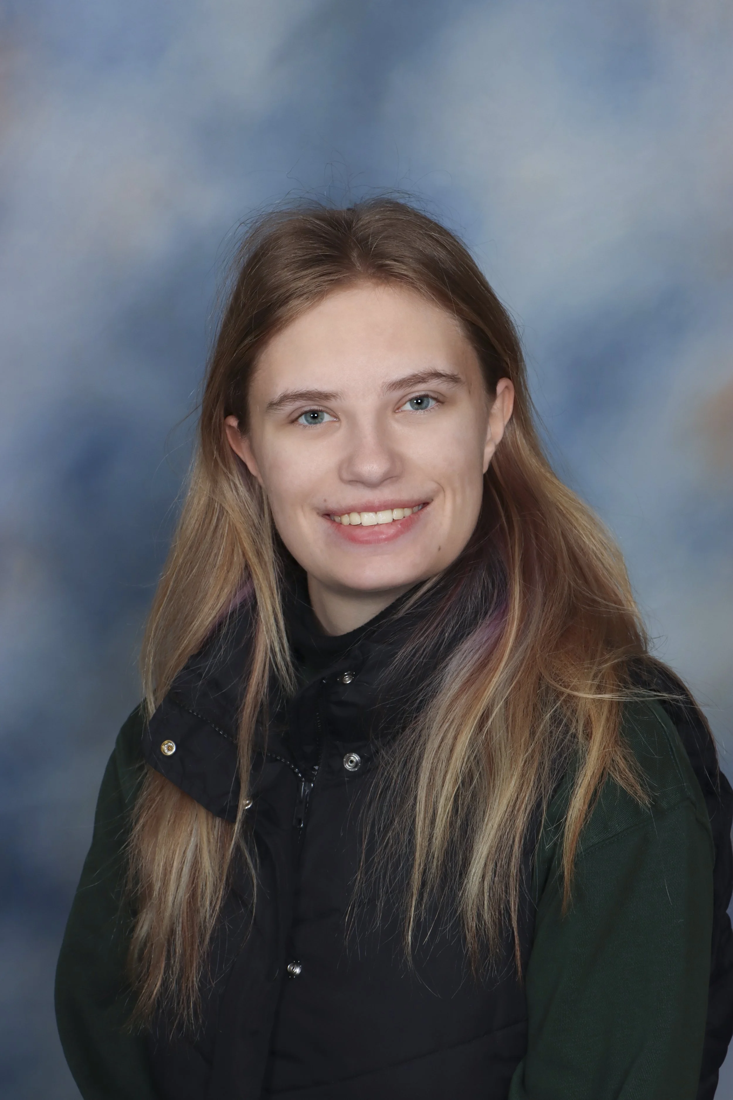 A young woman with blonde hair, wearing red lipstick and a black sleeveless top, smiling at the camera. She has a tattoo on her upper left arm and is wearing a black bandana with white musical notes.
