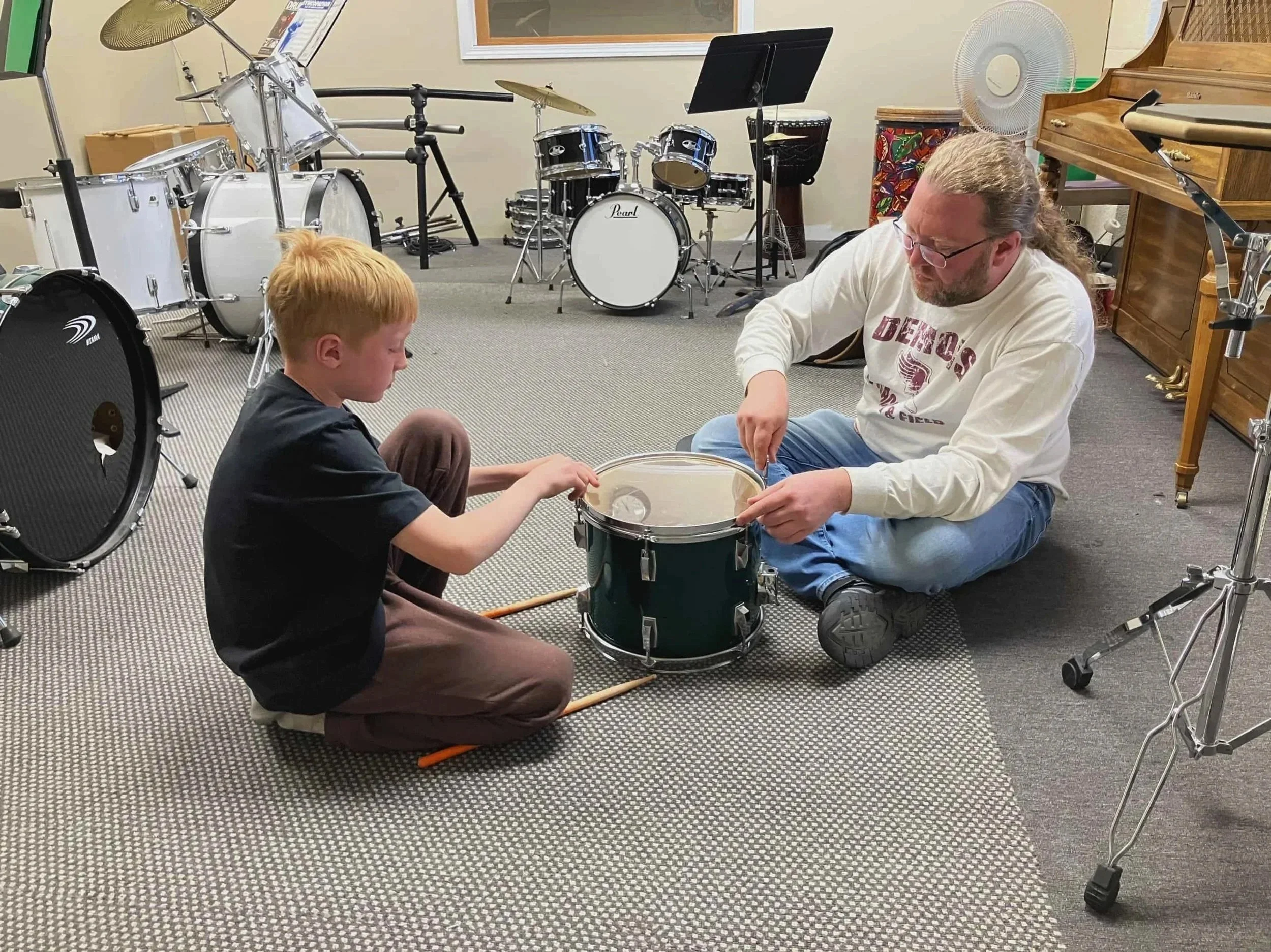 A man and a young boy sitting on the floor in a music room, working together on a drum set. The room has multiple drums, a keyboard, and music stands.