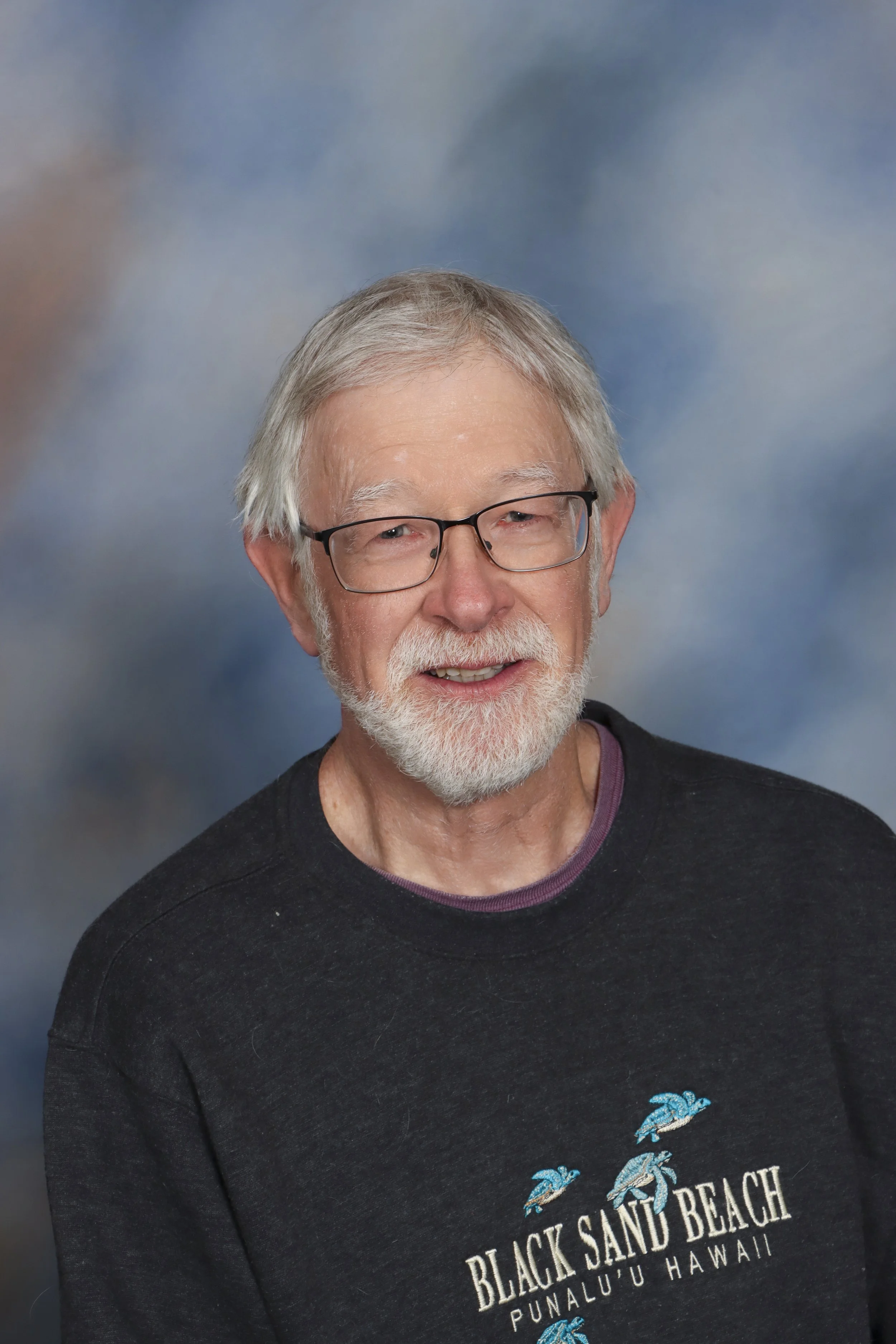 A man with white hair, beard, and glasses wearing a red checkered shirt, smiling against a gray background.