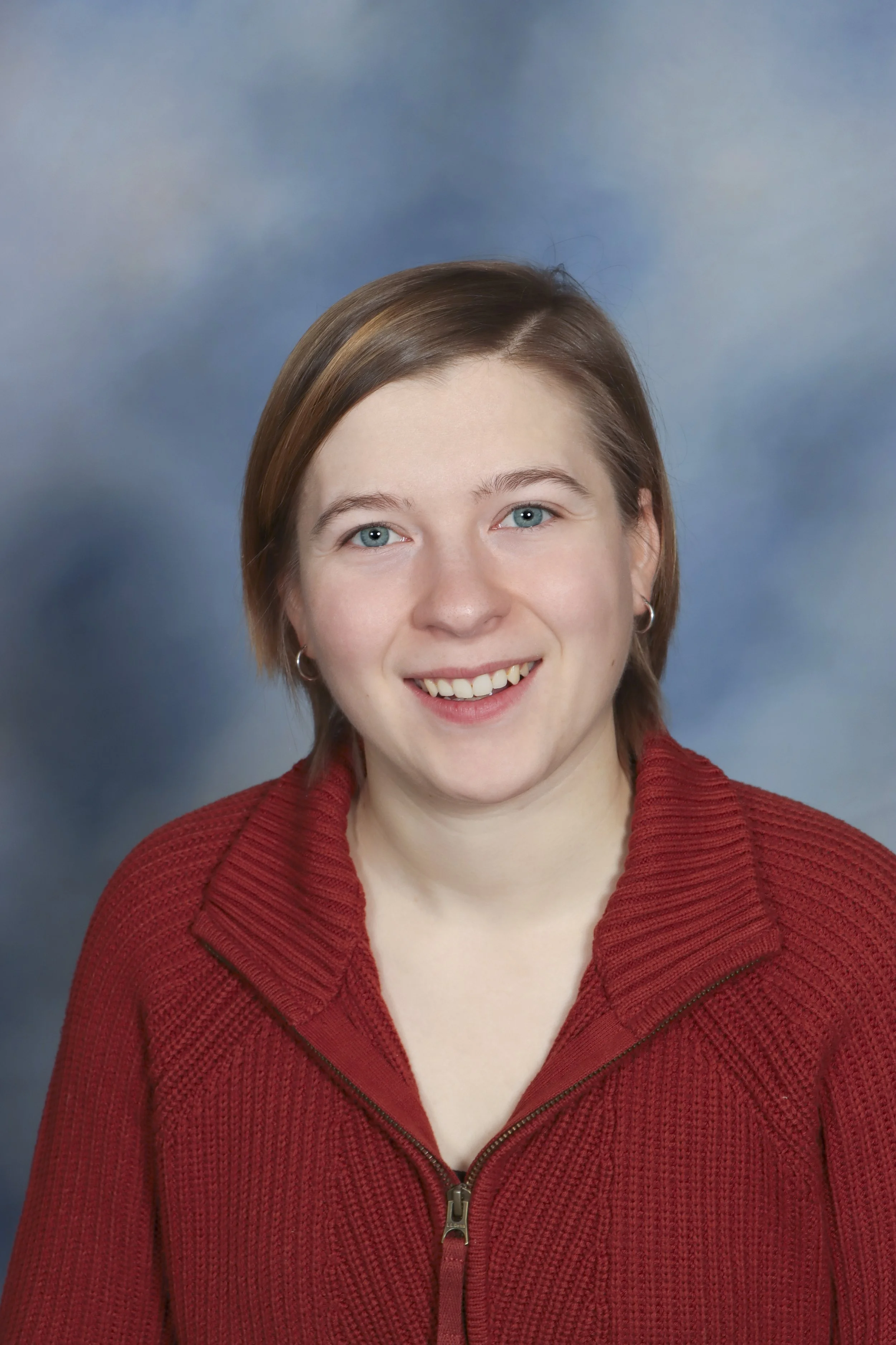 A young woman with light brown hair and blue eyes smiling, wearing a green jacket and a necklace with a pendant, standing in front of a light-colored wall.