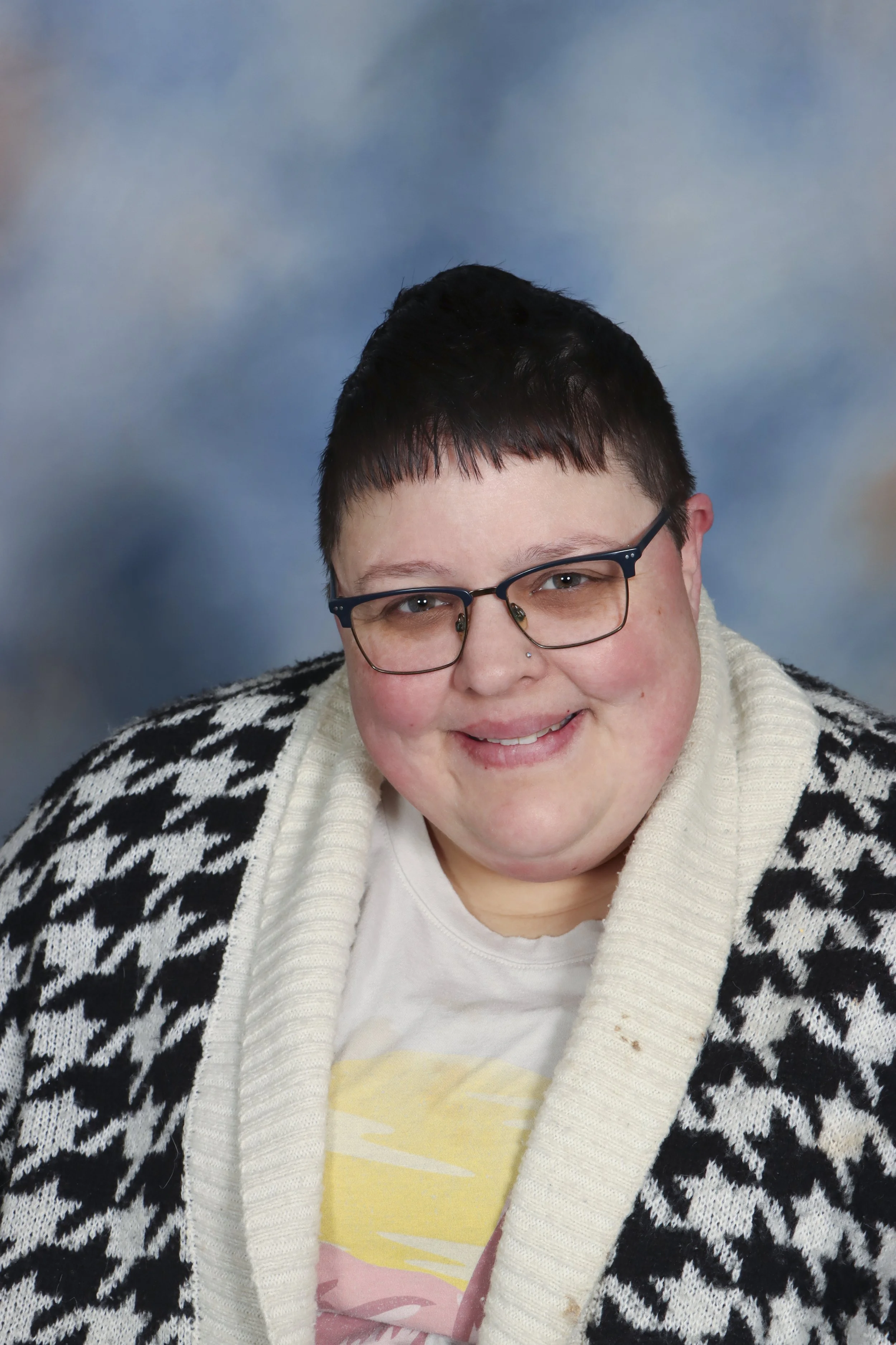 A smiling person with short dark hair and glasses, wearing a black sleeveless shirt, standing in front of a colorful abstract mural.