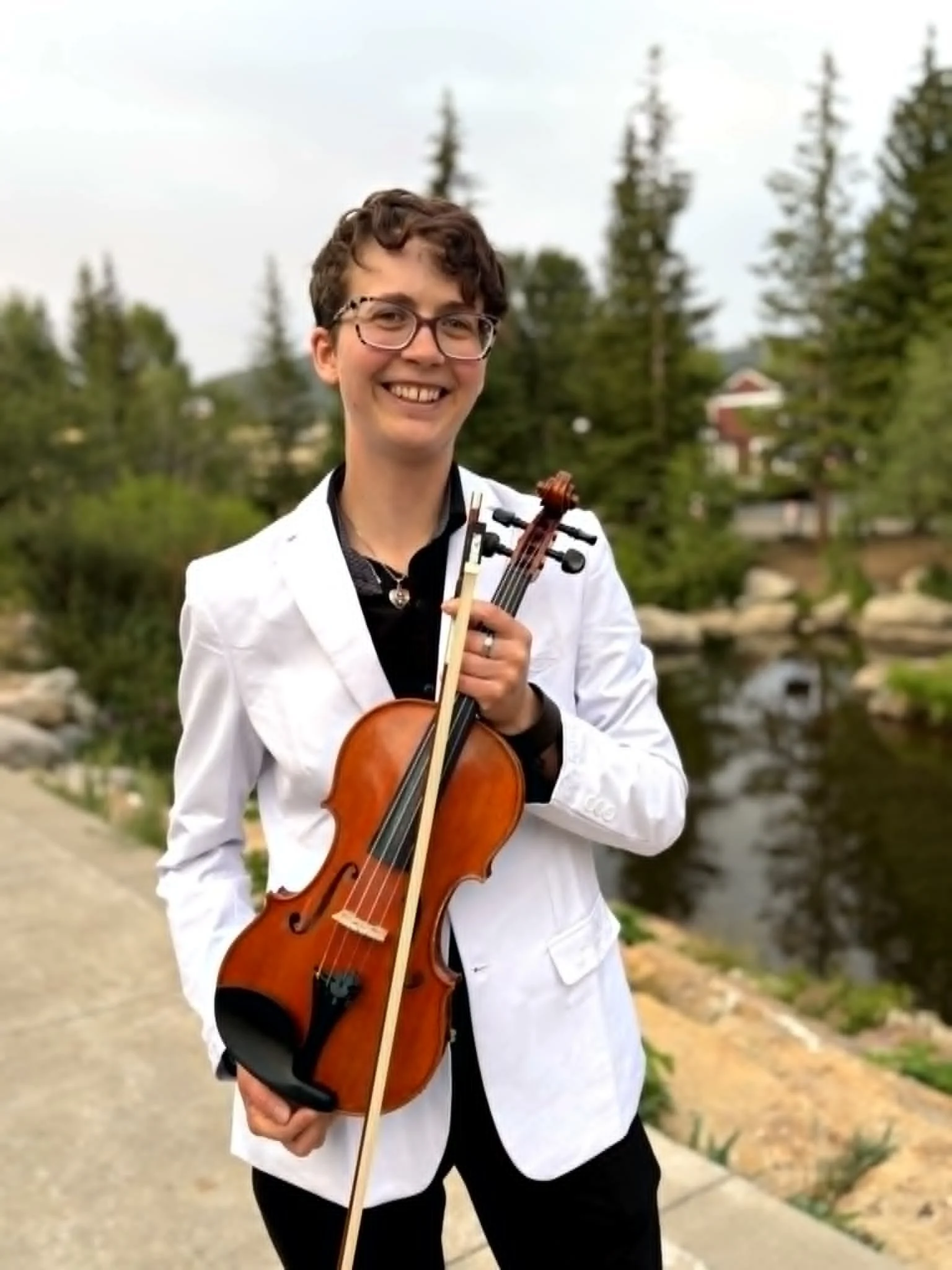 A young person with curly hair and glasses holding a violin while sitting at a piano.