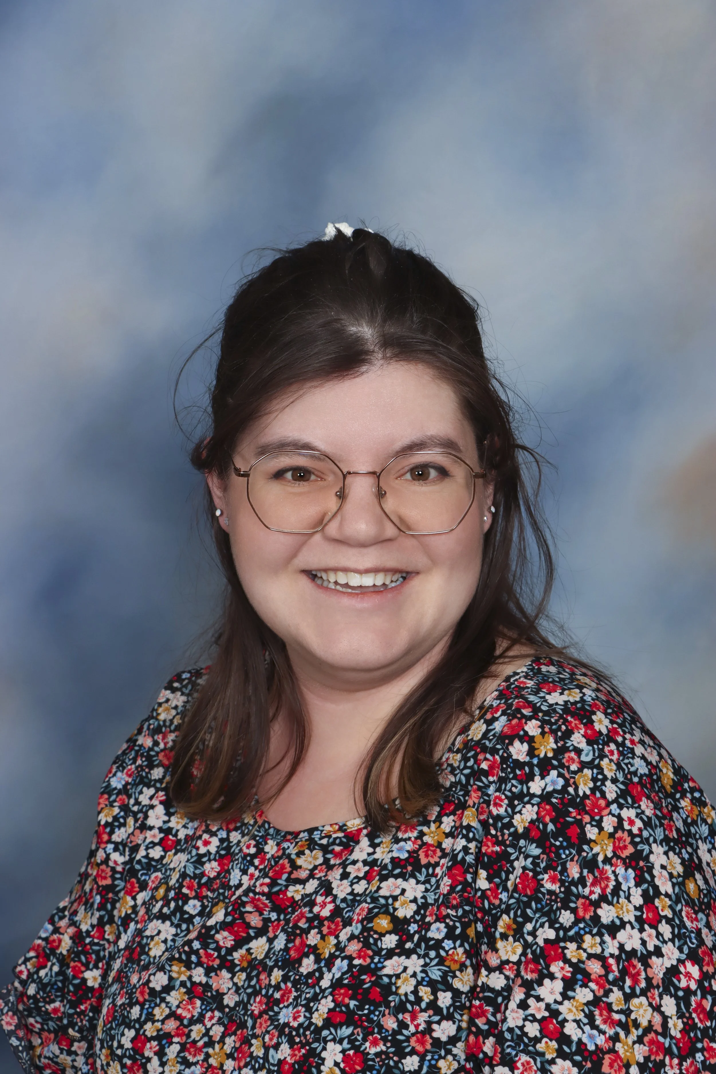 A woman with shoulder-length brown hair and glasses, smiling and wearing a brown top with a decorative necklace, in a room with a beige tiled wall.