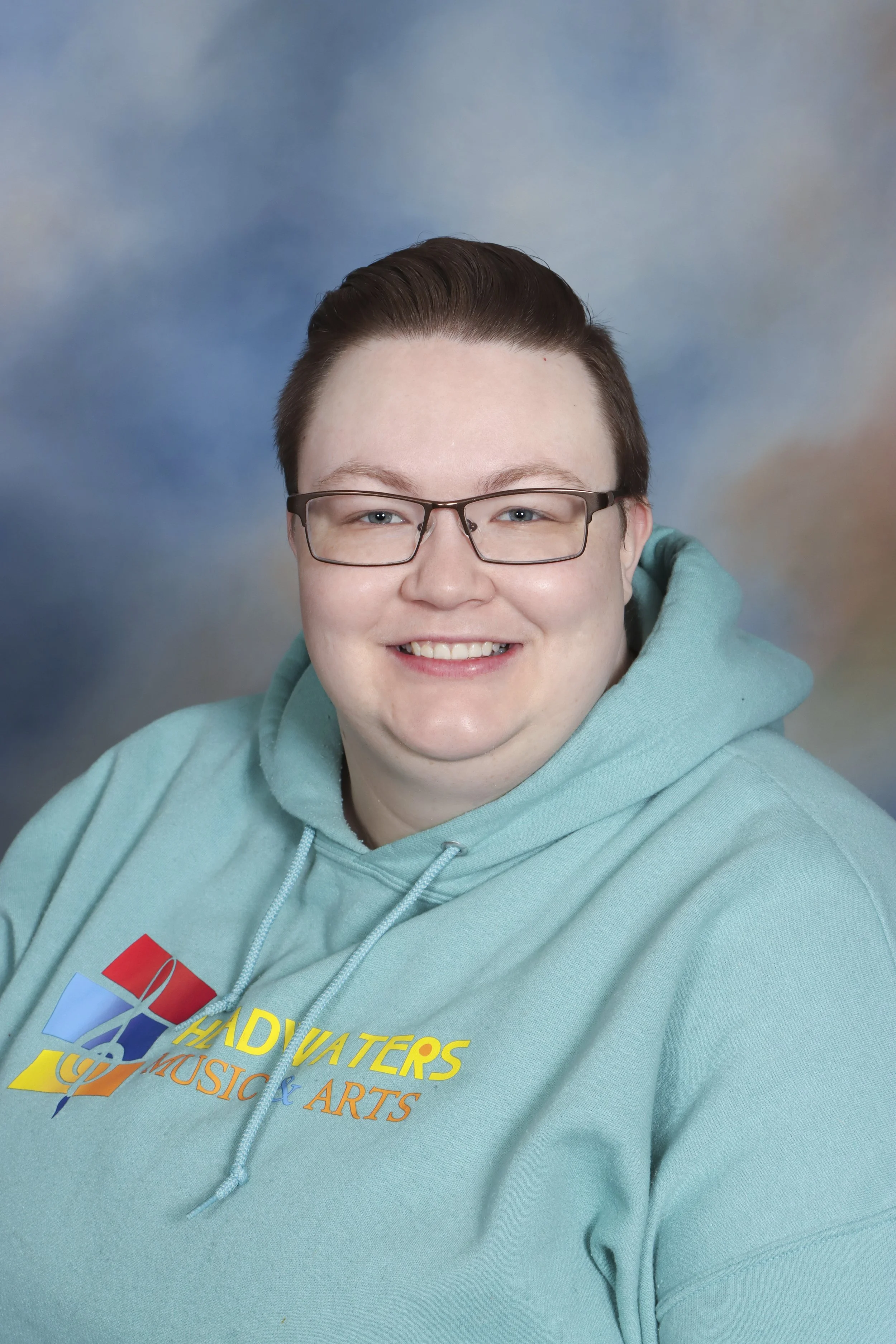 A person with short brown hair and glasses smiling at the camera, wearing a dark shirt, against a wooden background.