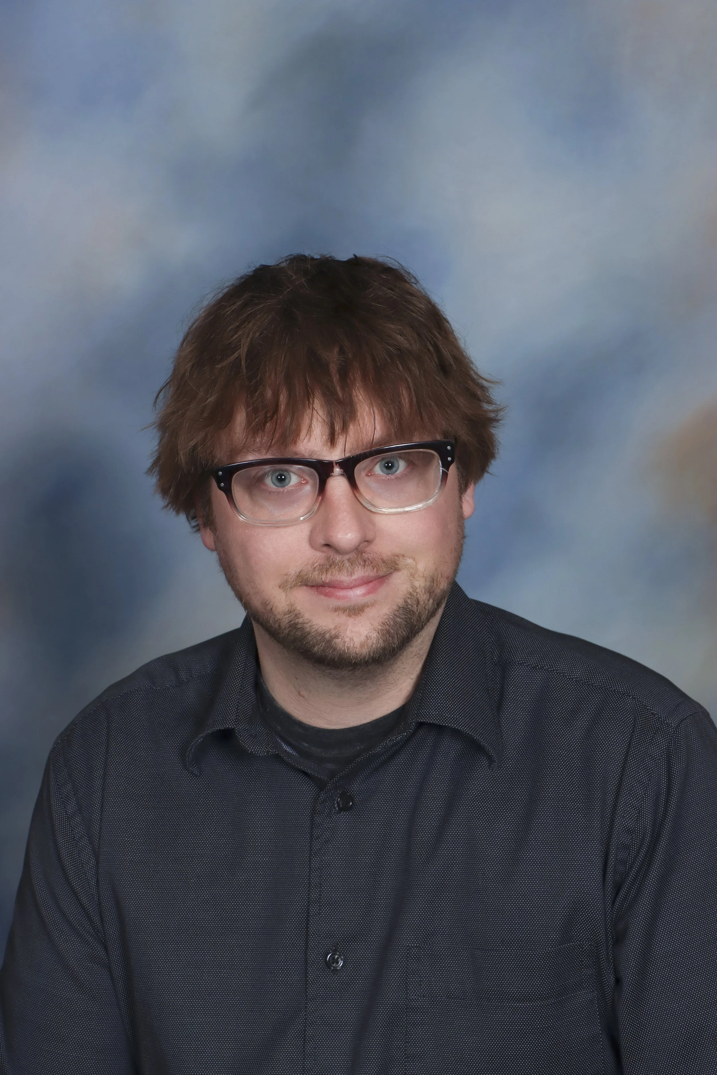 A person with glasses, a beard, and long hair wearing a maroon beanie hat and a gray jacket, standing in front of a circular decorative window with red brick-like framing and colorful lights inside.