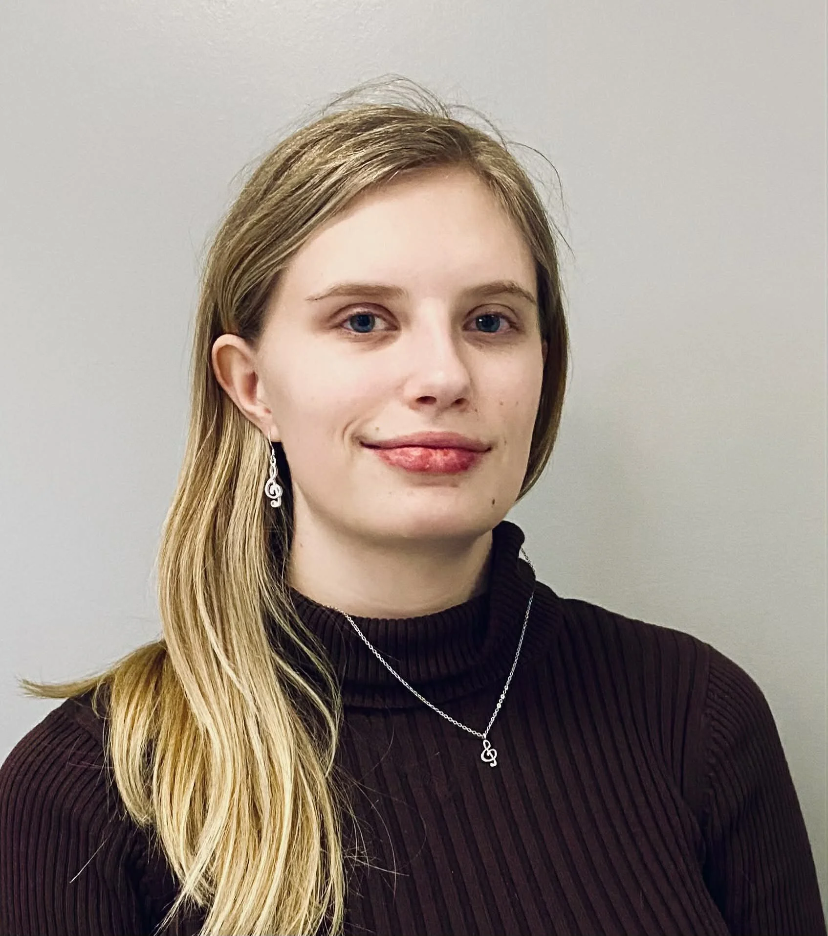 A young woman with blonde hair, wearing red lipstick and a black sleeveless top, smiling at the camera. She has a tattoo on her upper left arm and is wearing a black bandana with white musical notes.