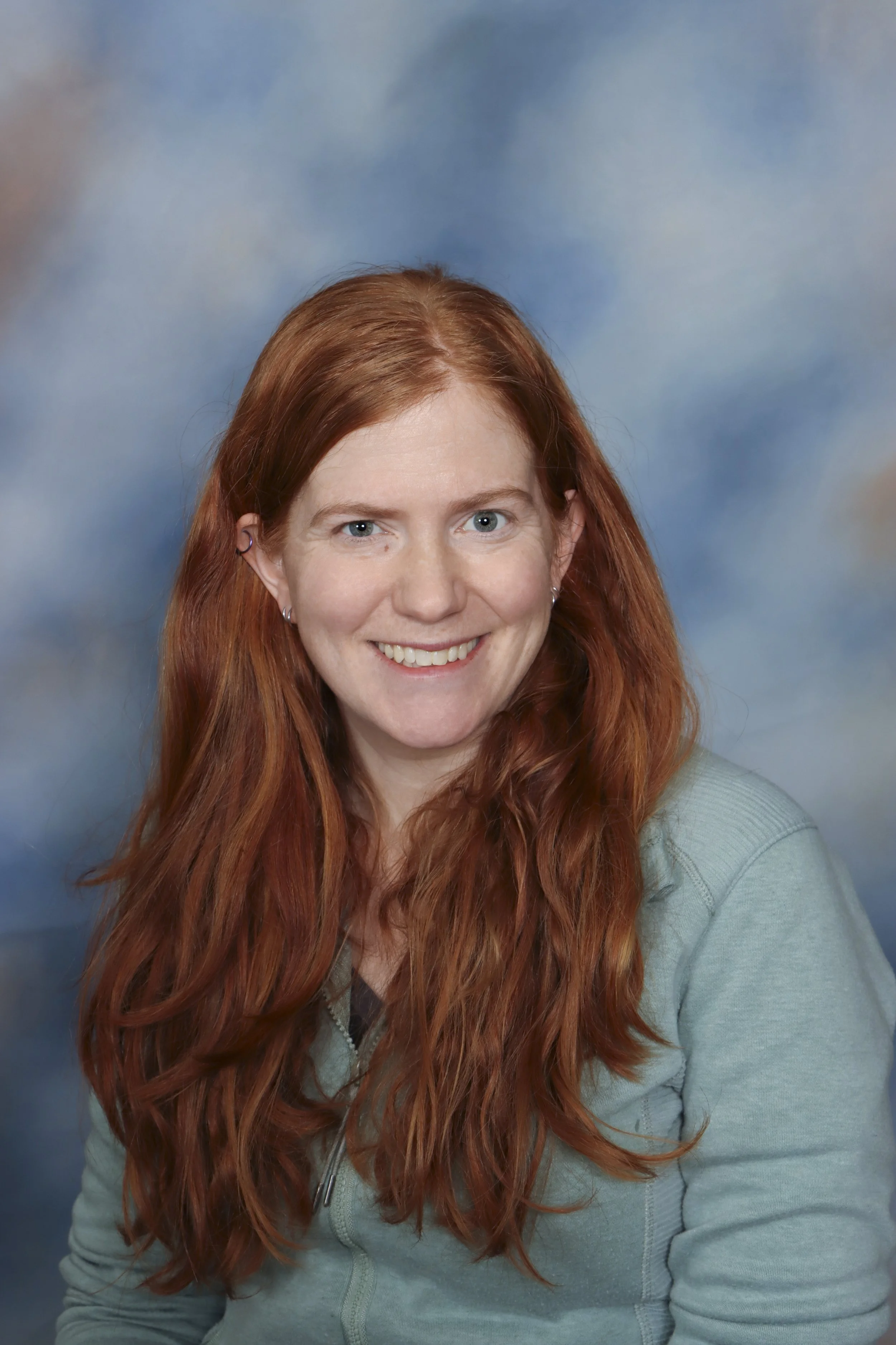 A woman with red hair smiling in front of a grand piano inside a room with wooden paneling and windows.