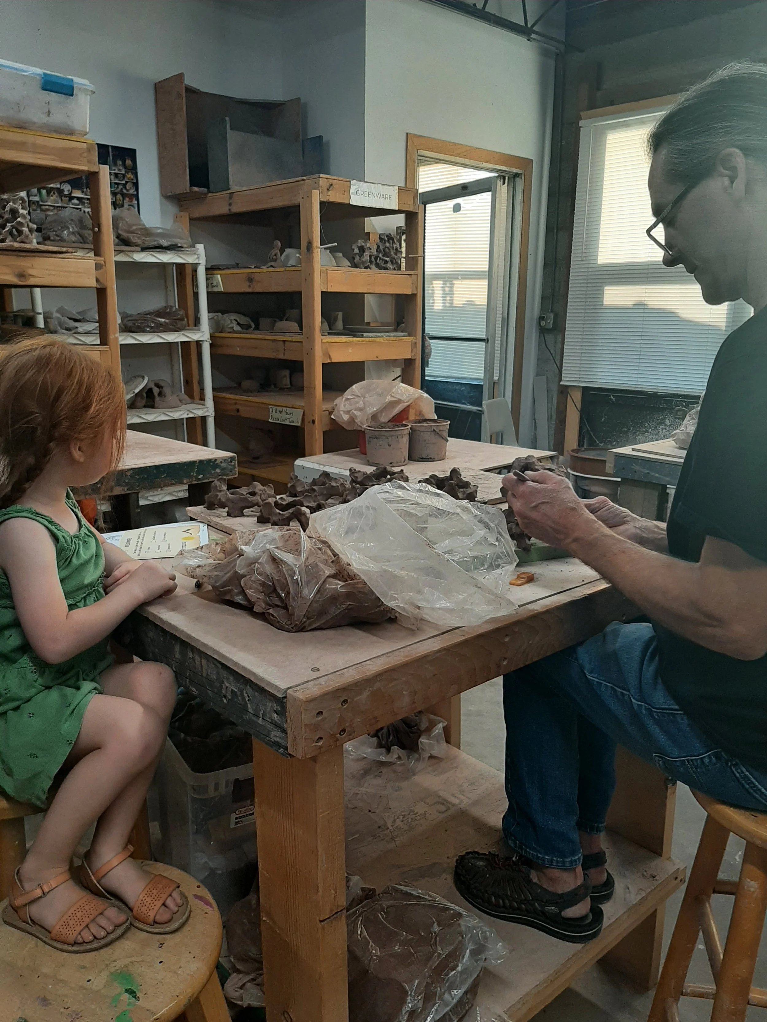 A young girl and an adult sit at a pottery table, shaping clay together in a studio filled with shelves of ceramic pieces.