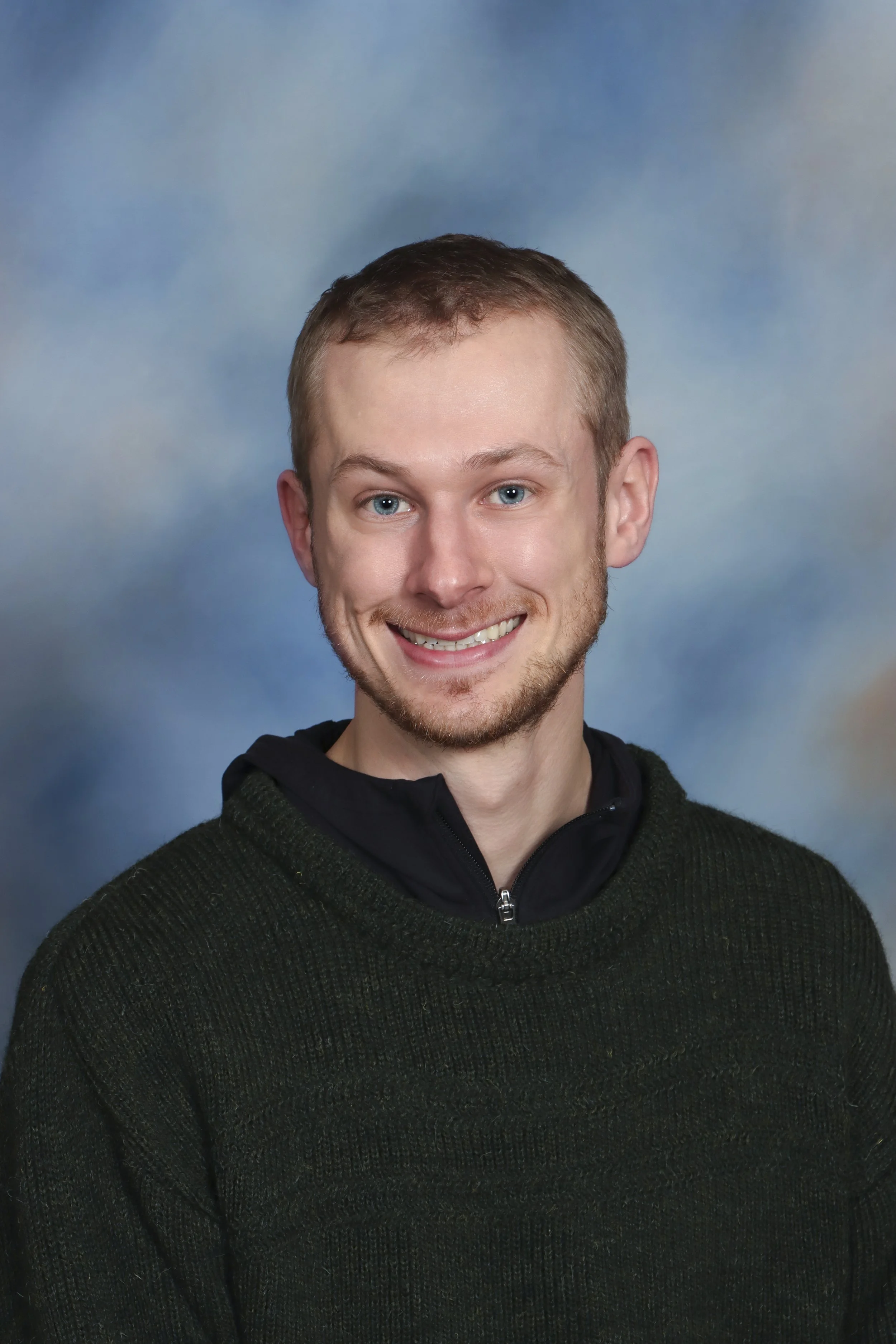 A young man with short light brown hair, blue eyes, and a smile, wearing a gray button-up shirt, standing in front of a wooden wall.
