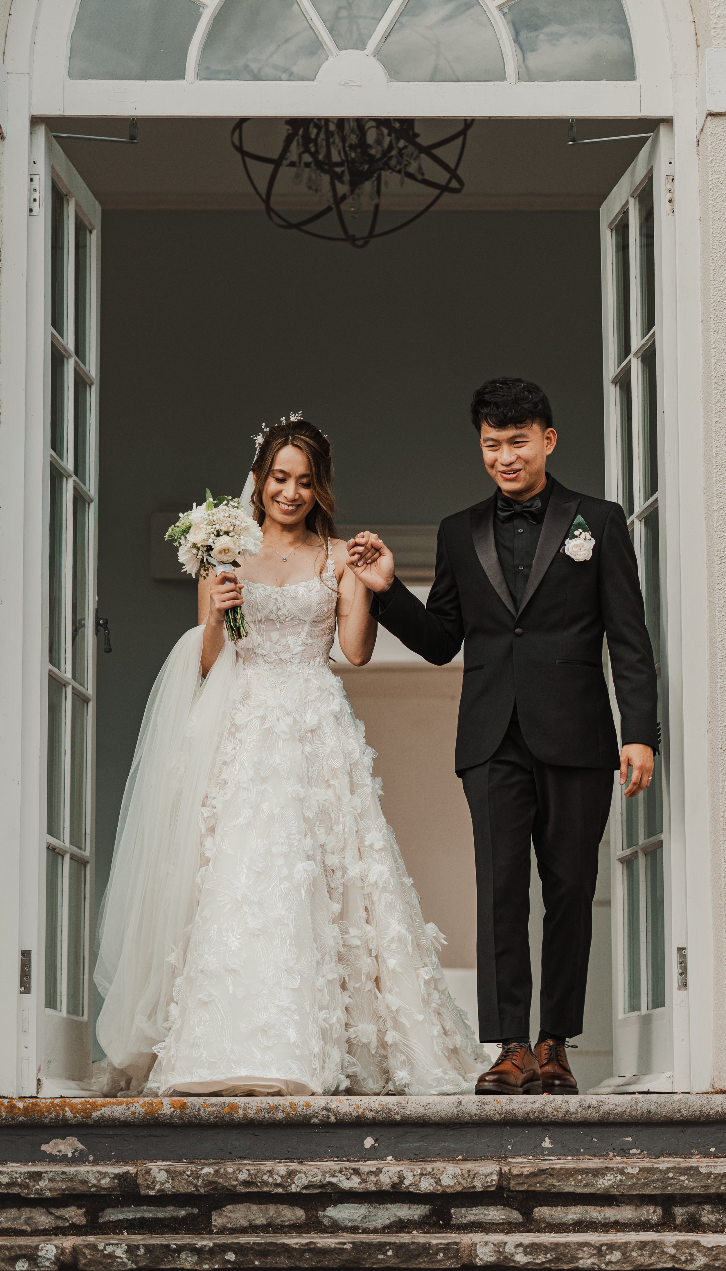 A bride and groom holding hands and smiling at each other outside in front of a large white building with multiple windows, with greenery and a rusted decorative stone urn in the foreground.
