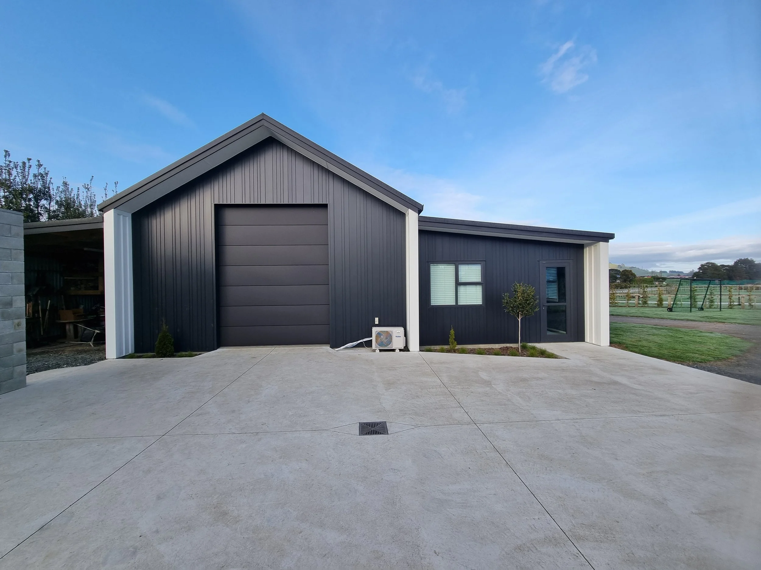 Modern black house with a garage door, small window, and a glass door, surrounded by a concrete driveway and some small plants, with a fence and play area in the background under a clear blue sky.