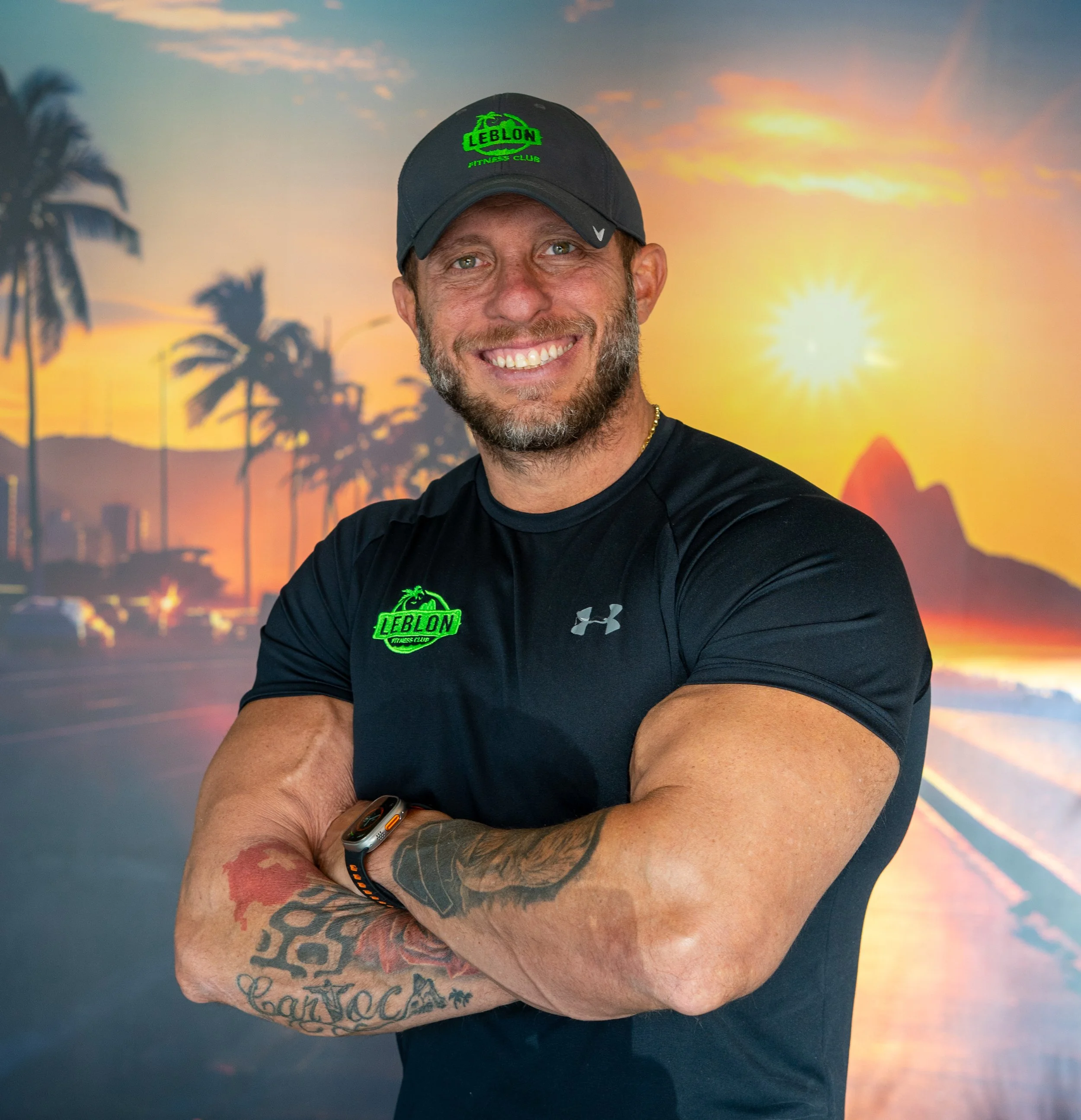 A smiling man with tattoos on his arms wearing a black athletic shirt and a black cap with green logo, standing with arms crossed in front of a tropical sunset backdrop with palm trees and a volcano.