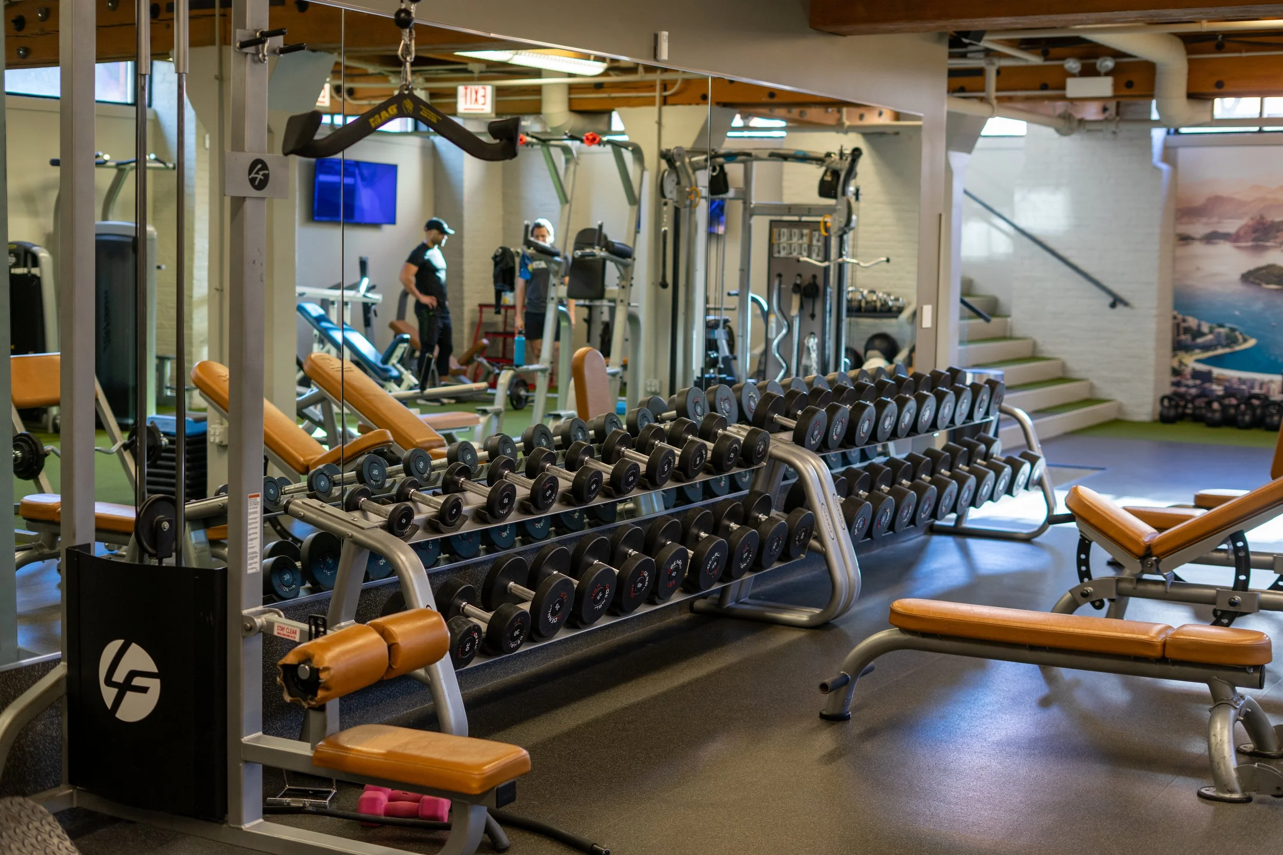 A gym with various weightlifting equipment including dumbbells on racks, benches, and machines, with people working out in the background.