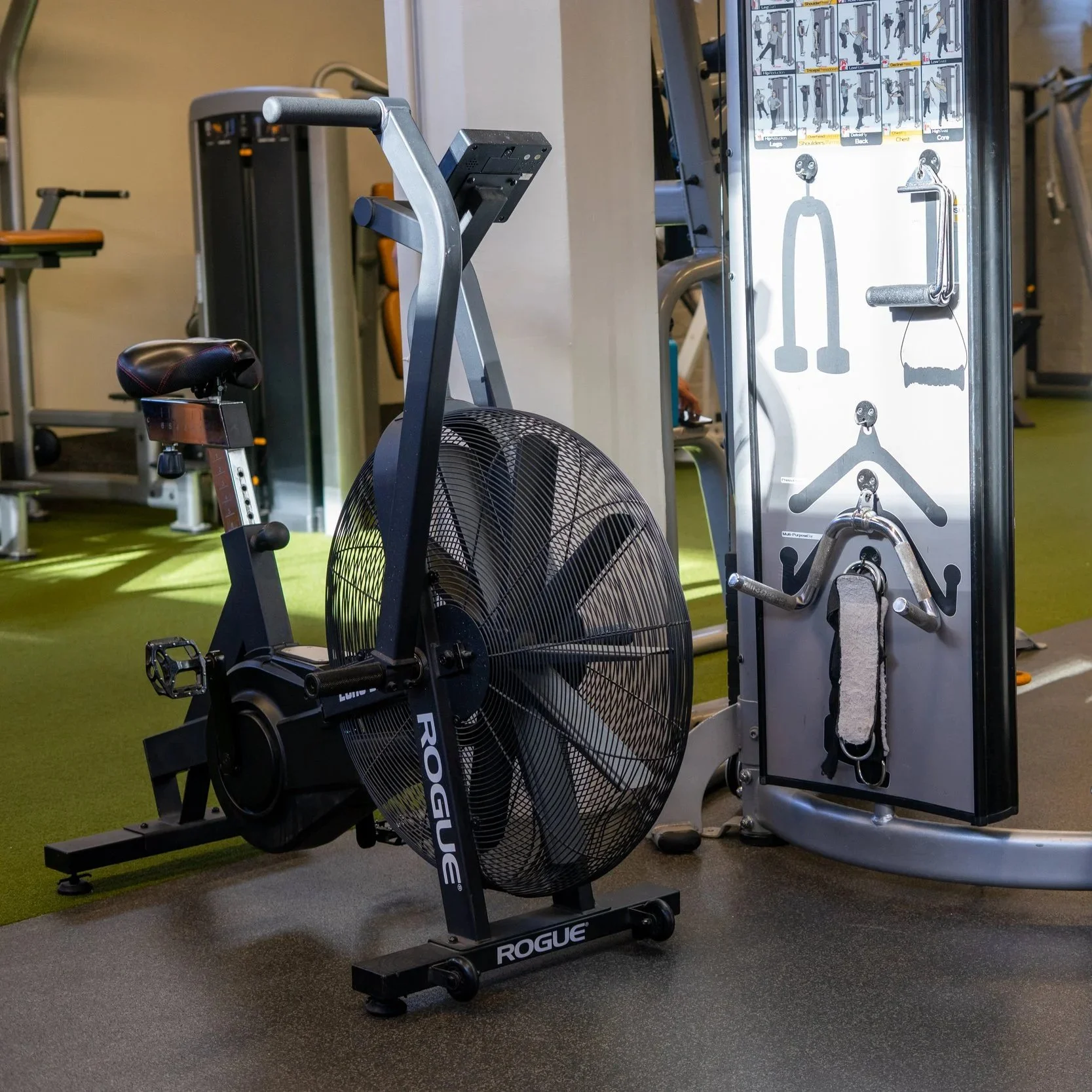 Indoor gym room featuring a Rogue fan bike, with exercise instructions on a nearby poster, and various gym equipment in the background.