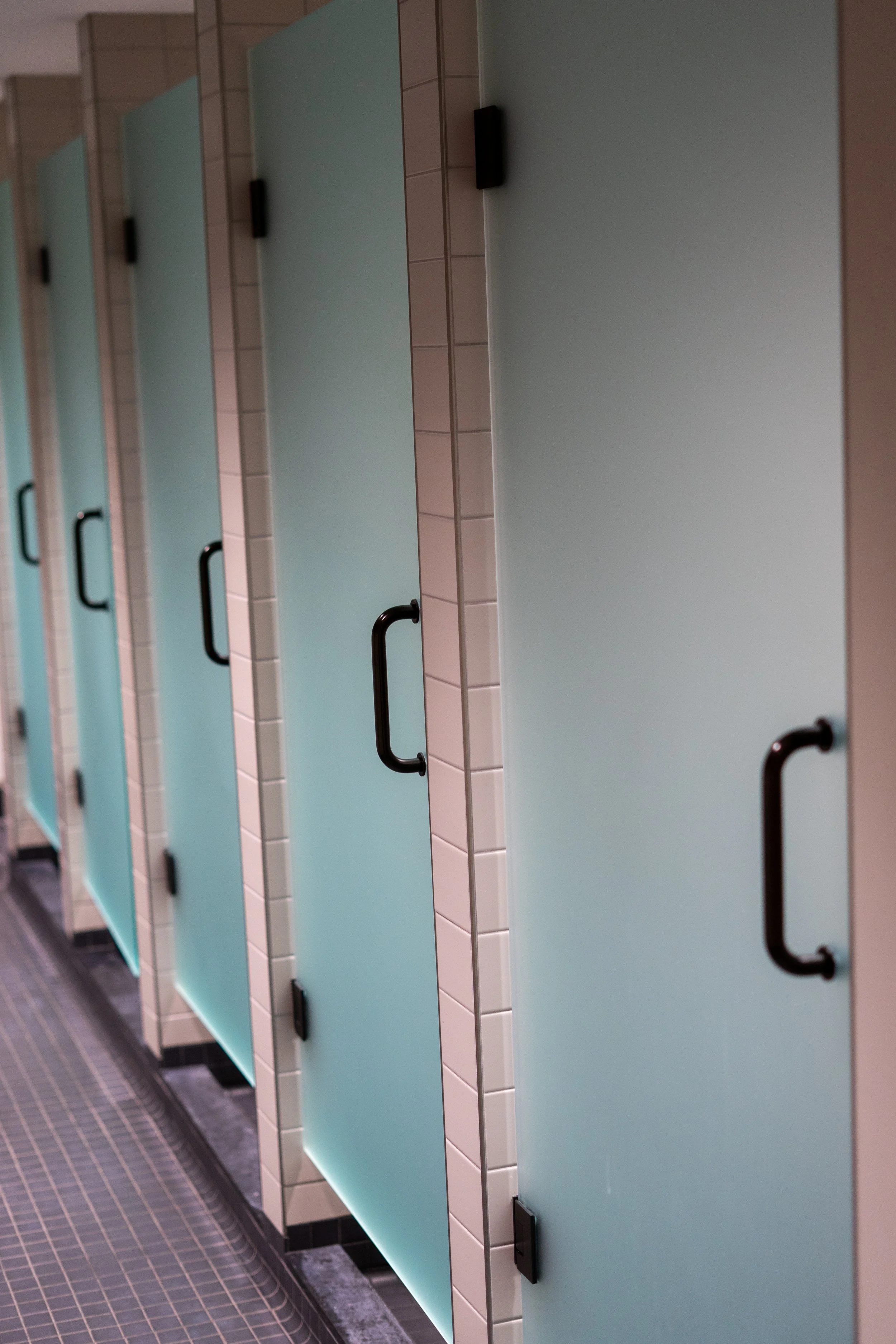 A row of teal bathroom stalls with black handles, beige tiled walls, and a dark tiled floor.