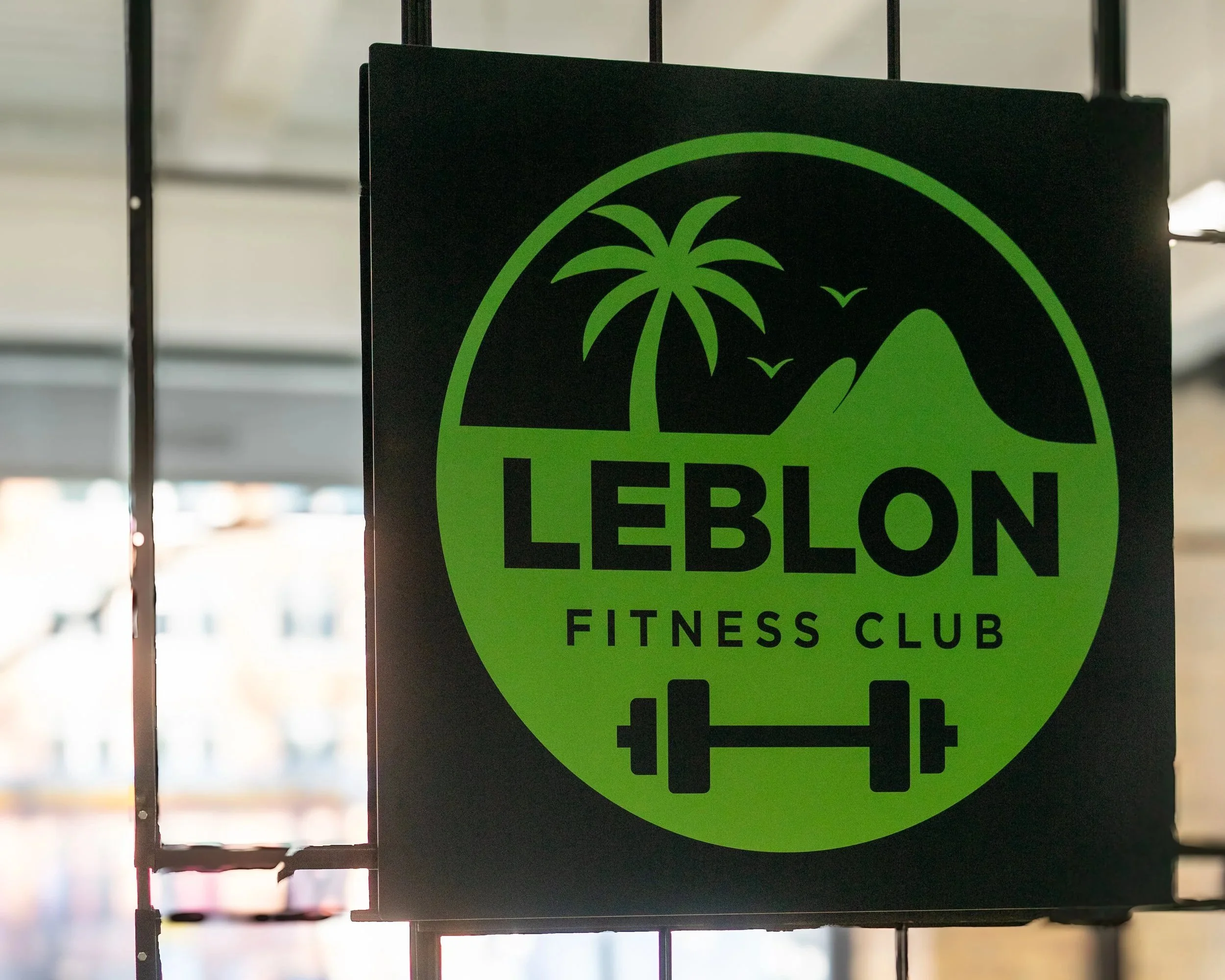 Sign for Leblon Fitness Club featuring a palm tree, mountains, birds, and a barbell in green and black colors.