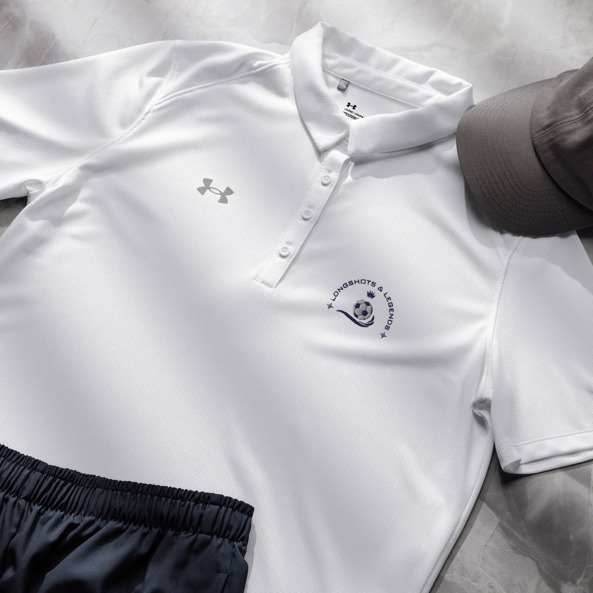 Custom embroidered women’s Under Armour® collared polo shirt with soccer logo in white. Celebrate with the Longshots and Legends collection of athletic wear.