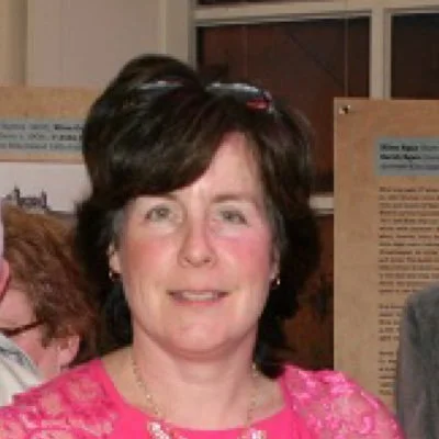 A woman with dark brown hair, wearing a pink lace top, smiling, standing indoors near a window and a brown poster board.
