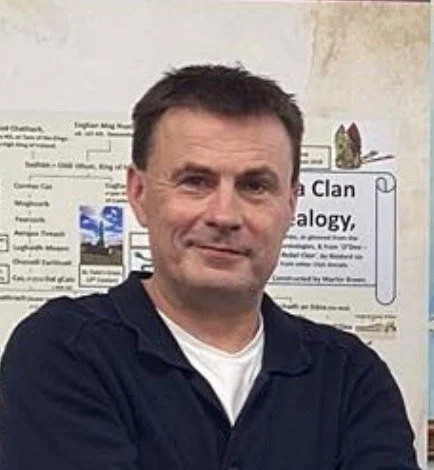 A man with short dark hair and a slight smile, standing in front of a bulletin board with various posters and papers, wearing a dark jacket over a white shirt.