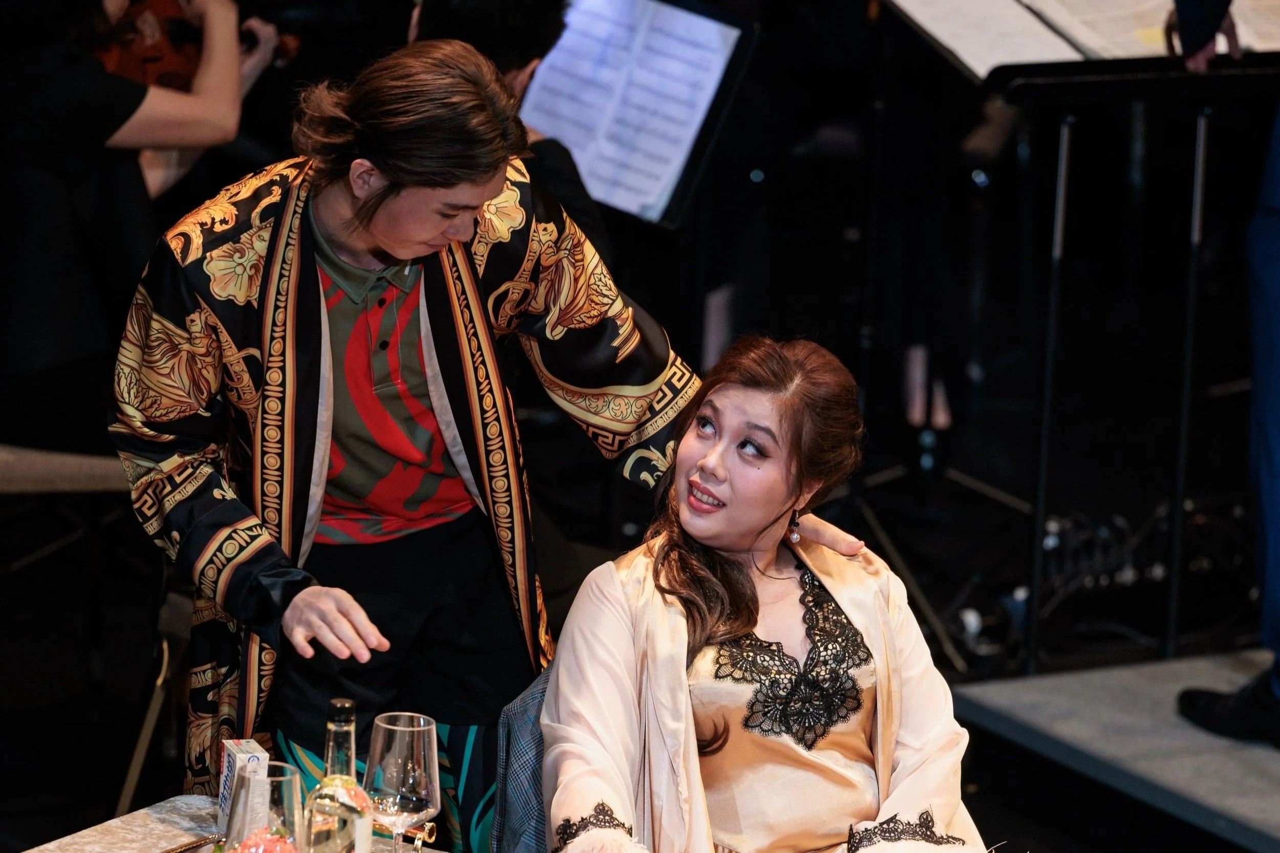 Two performers engaged in conversation. One with dark hair in a stylish patterned jacket leans over a seated woman with reddish-brown hair who is looking up.