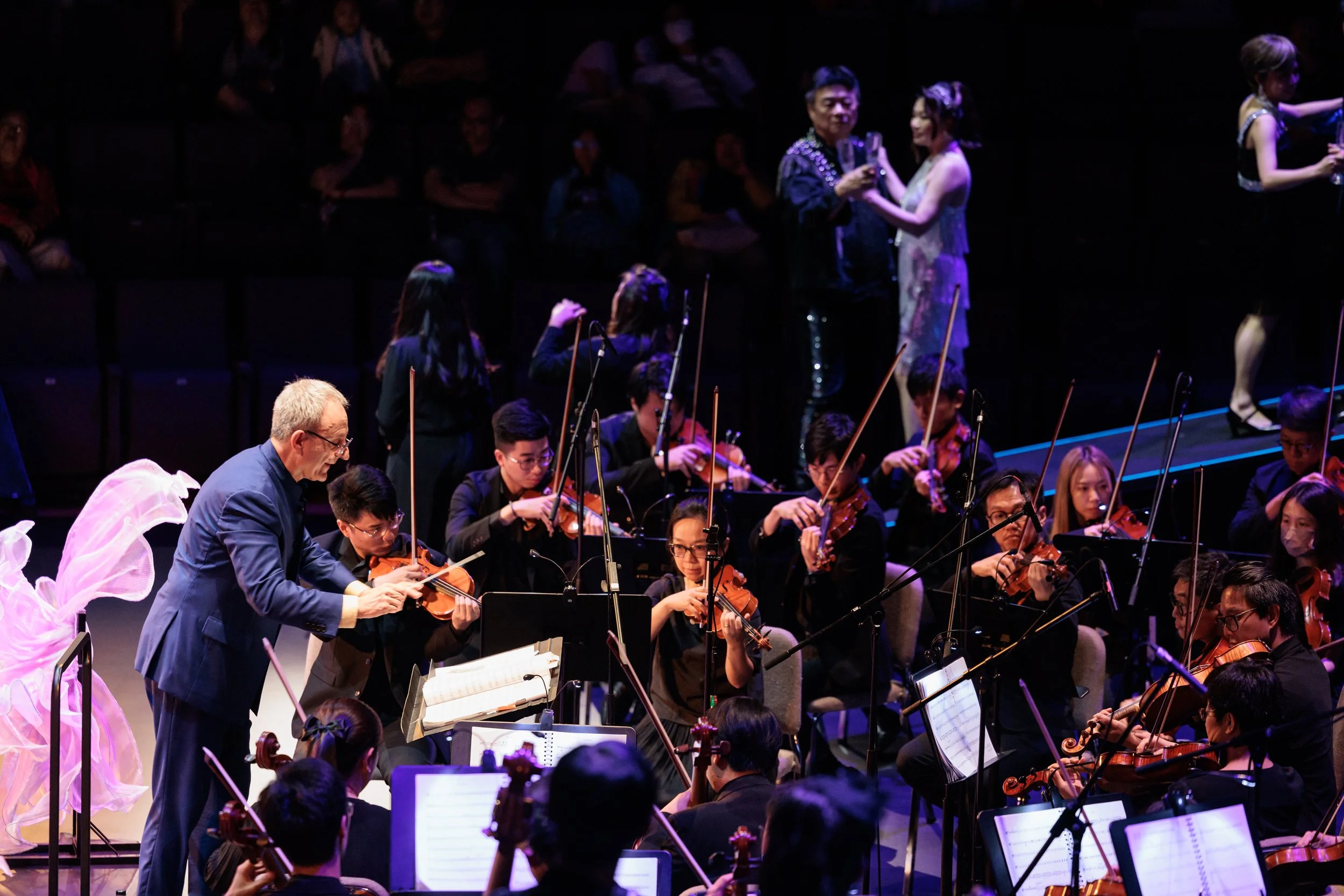 An orchestra performing on stage with a conductor at the front. The orchestra consists of violinists and other string players, with some audience members visible in the background. The stage is dimly lit with spotlights on the musicians.