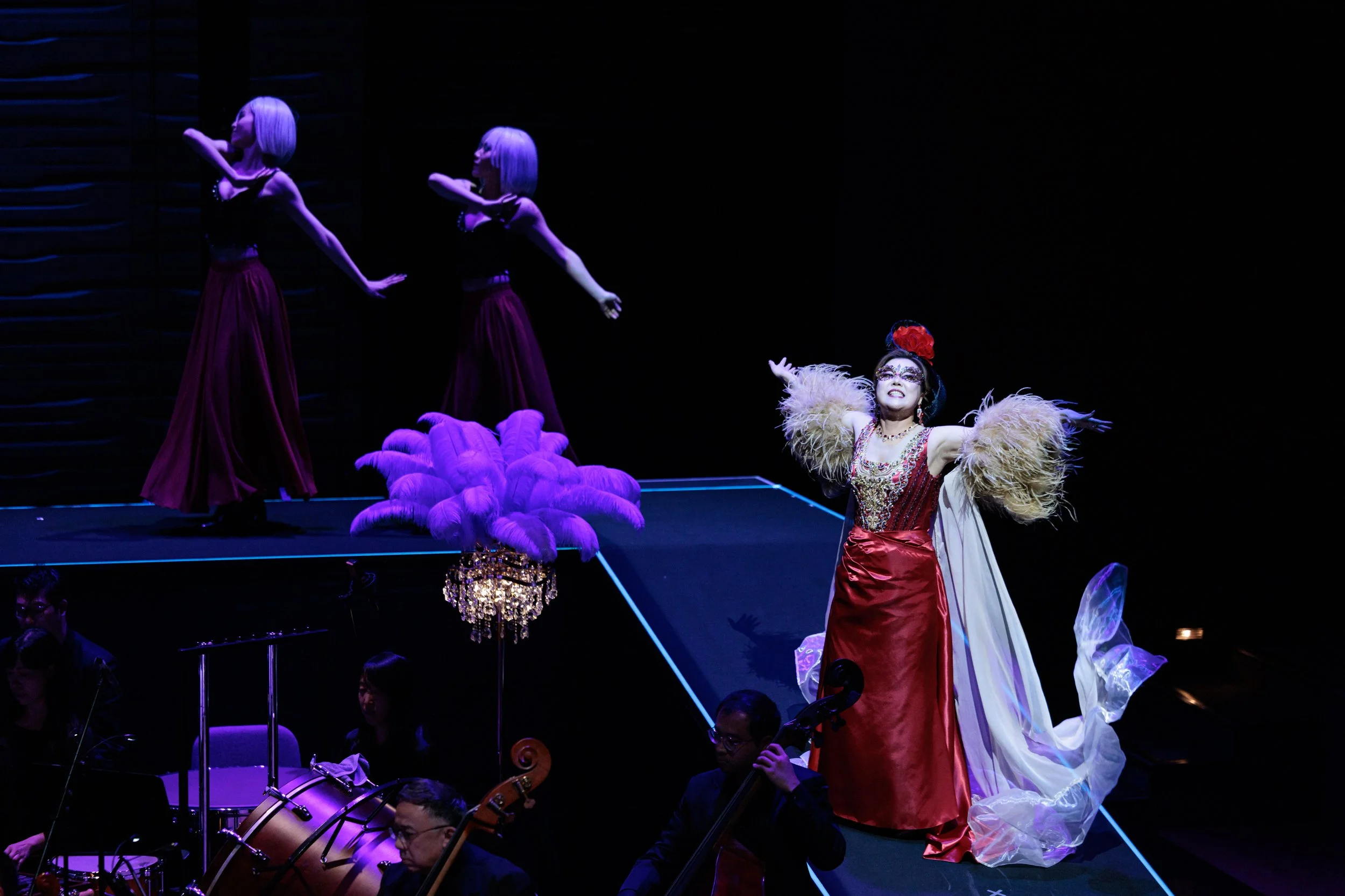 An opera stage with three female performers and musicians below. One performer in a red dress and a feathered headpiece is singing, while two others in purple dresses dance on an elevated platform.