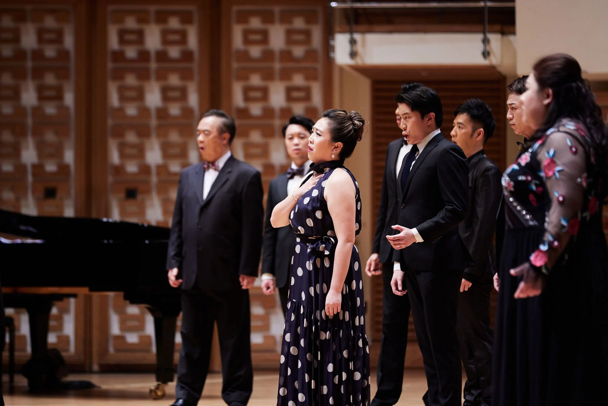 A group of seven singers standing in a line in on opera performance setting, some with hands over their hearts, with a grand piano on the left, in a room with wooden walls and floors.