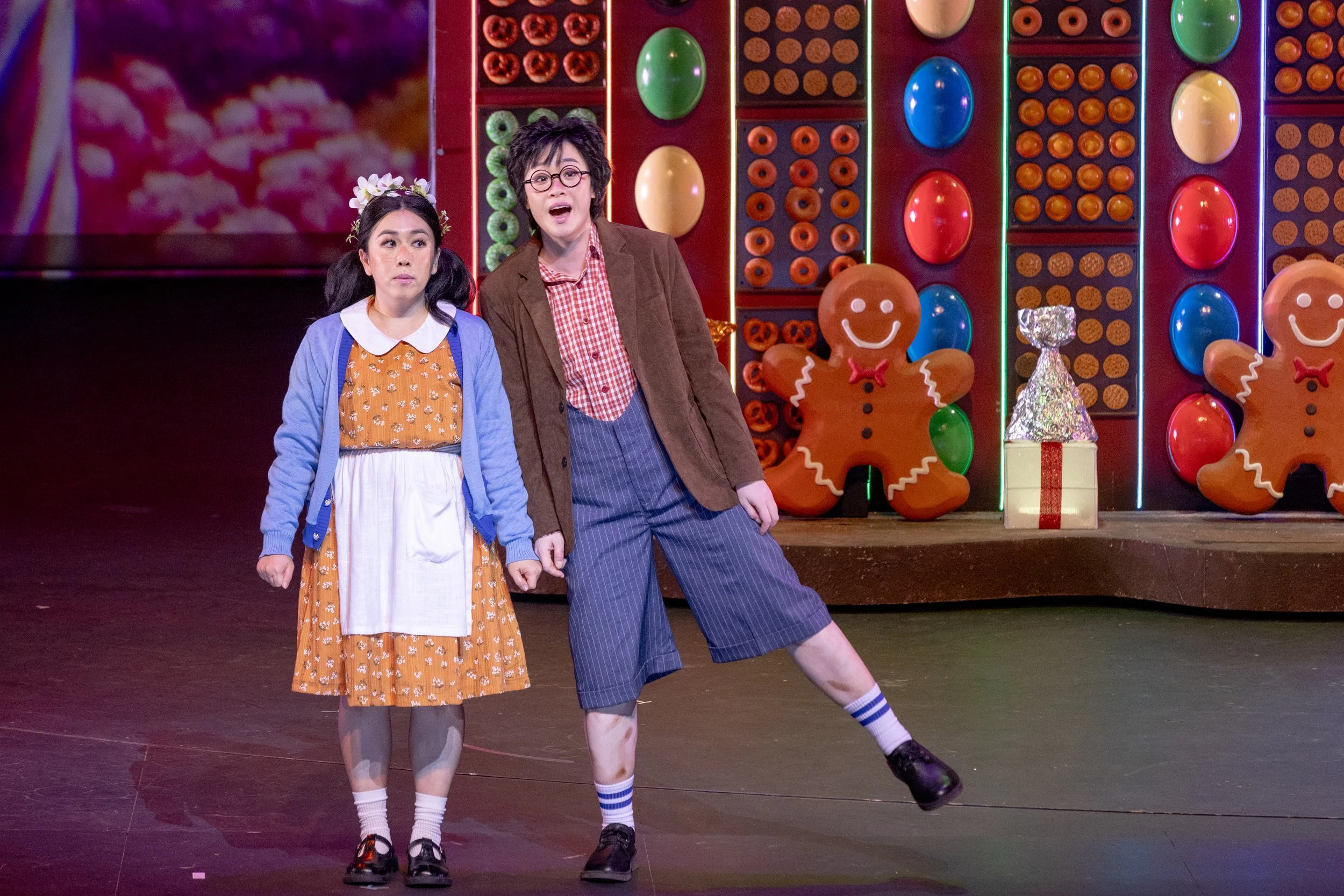 Two opera singers in colourful costumes performing on stage with Christmas gingerbread figures and wrapped gift decorations in the background.