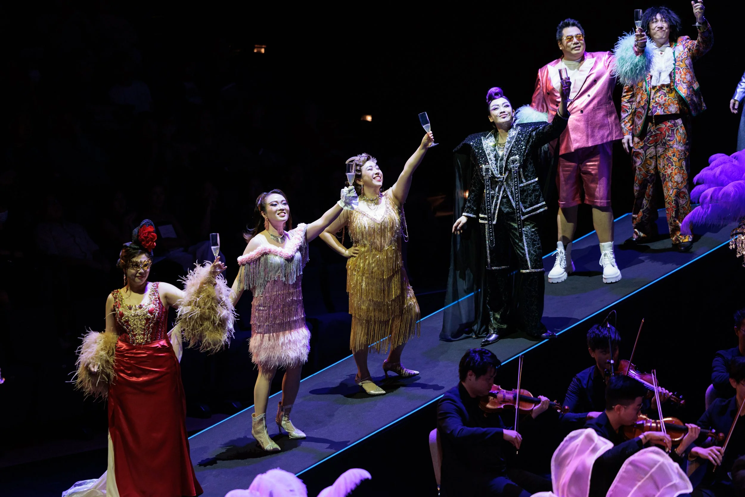The performers on stage with elegant and colorful costumes, holding glasses of champagne, and the orchestra playing music in the foreground.
