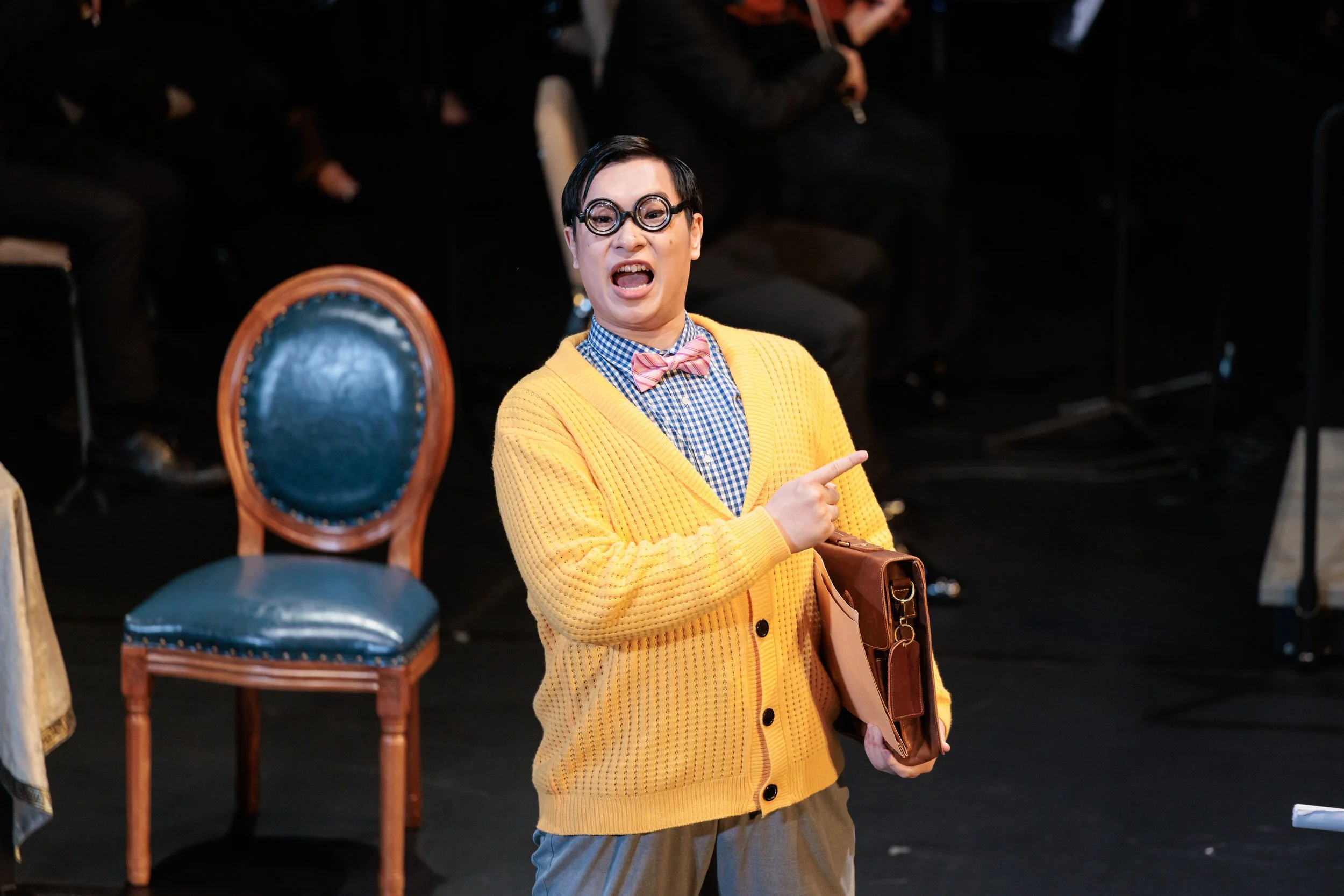 A person with short dark hair and round glasses, wearing a yellow cardigan, checkered shirt, pink bow tie, and khaki pants, pointing and singing passionately. They are holding a folder, with an empty blue chair in the background on a stage.