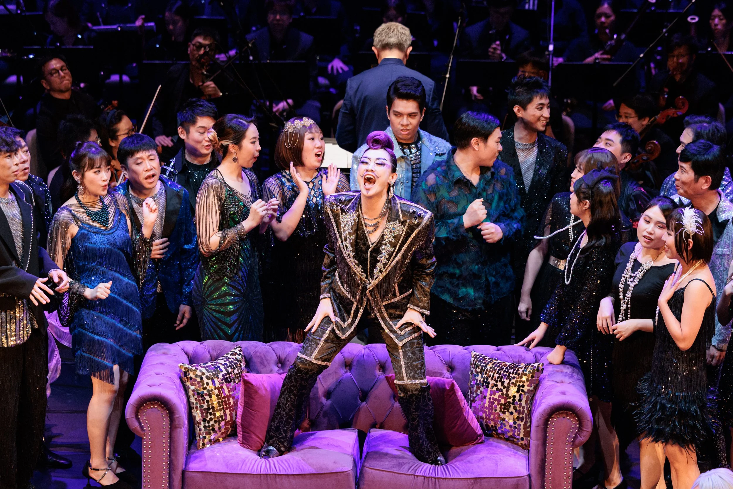 A lively opera stage scene with a group of performers dressed in glamorous, shiny costumes, surrounding a woman standing on her sofa, singing enthusiastically. The orchestra is visible in the background.