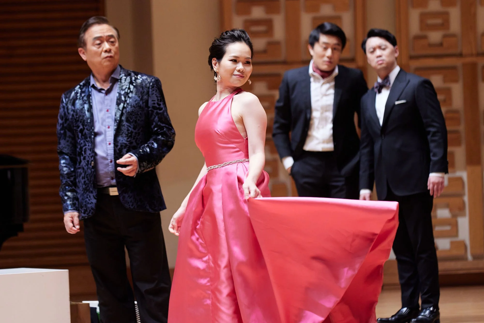 A woman in a pink gown holding up her dress while four men in suits stand nearby as part of an operatic scene.