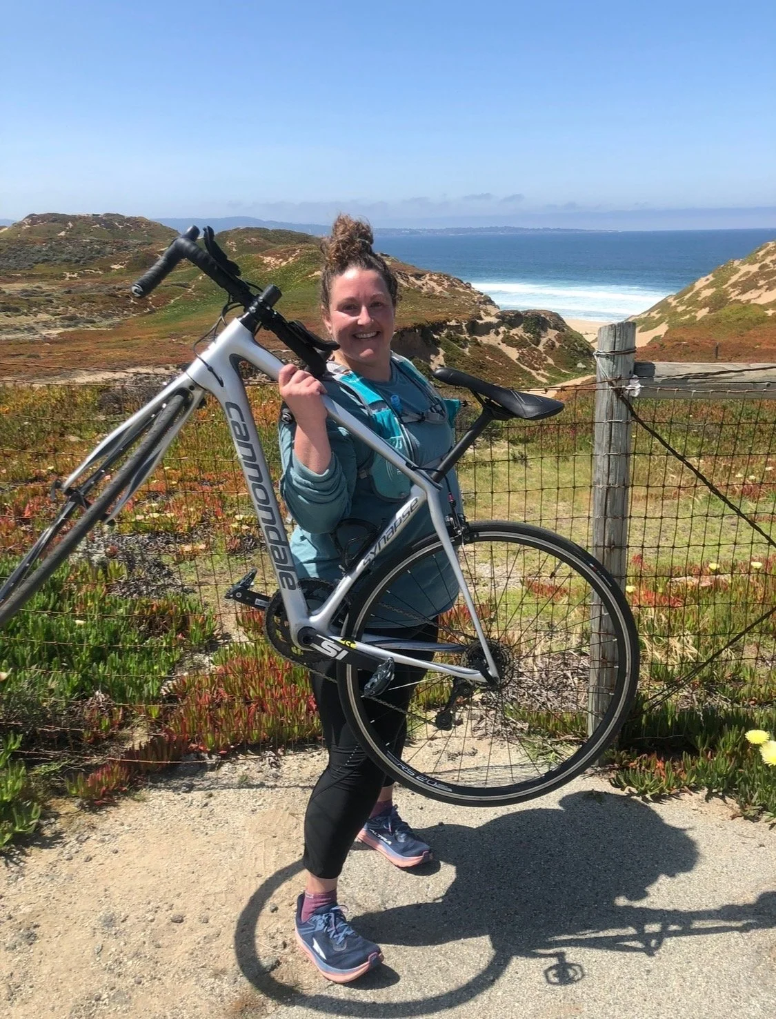 Picture of Hayley, a Well With Avril stress management and lifestyle client, holding her bike and posing against a backdrop of coastal path with ocean views