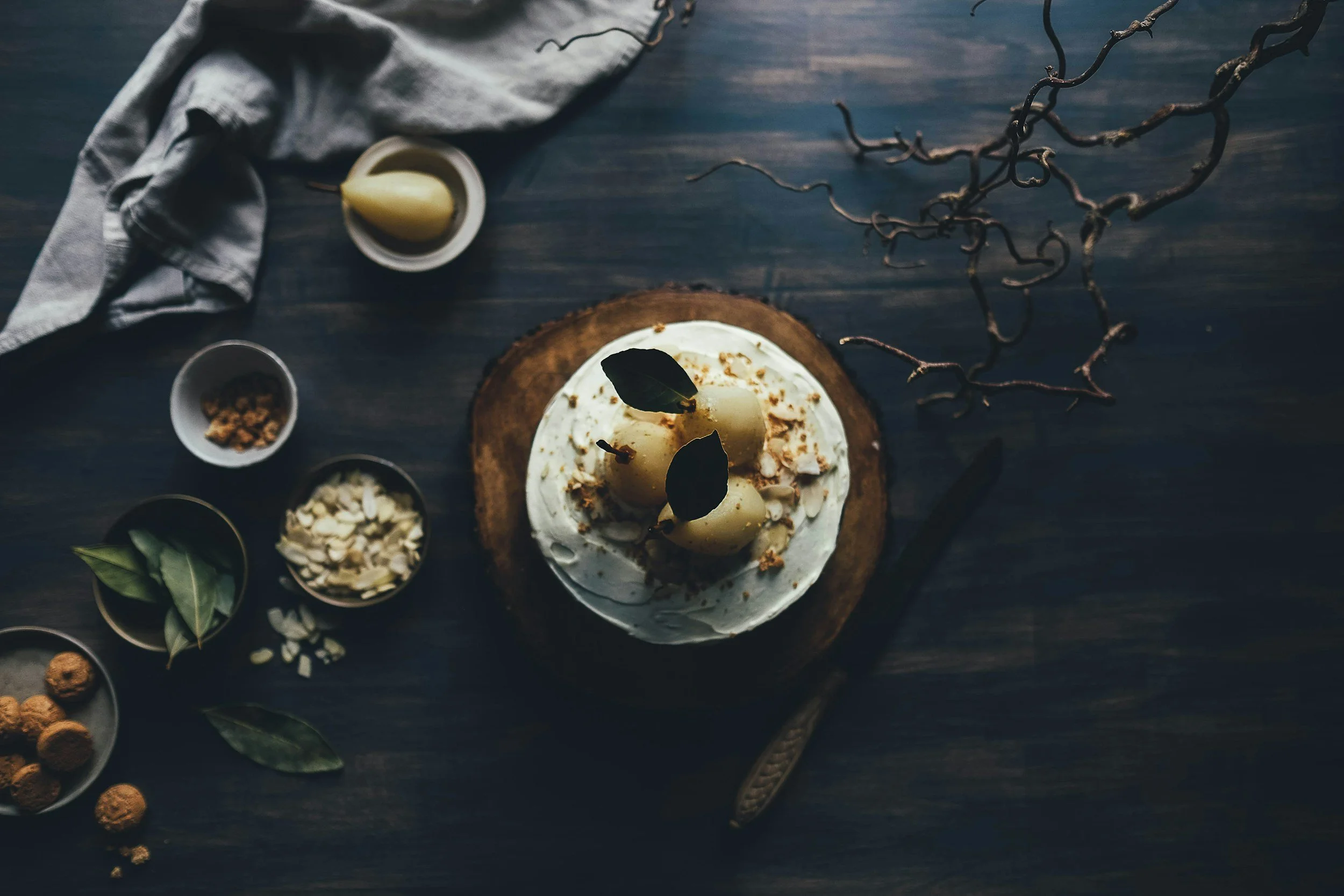 Overhead view of a cake with white frosting, topped with poached pears and leaves, surrounded by small bowls containing almonds, brown sugar, and bay leaves on a dark wooden table with a cloth napkin and decorative twigs.