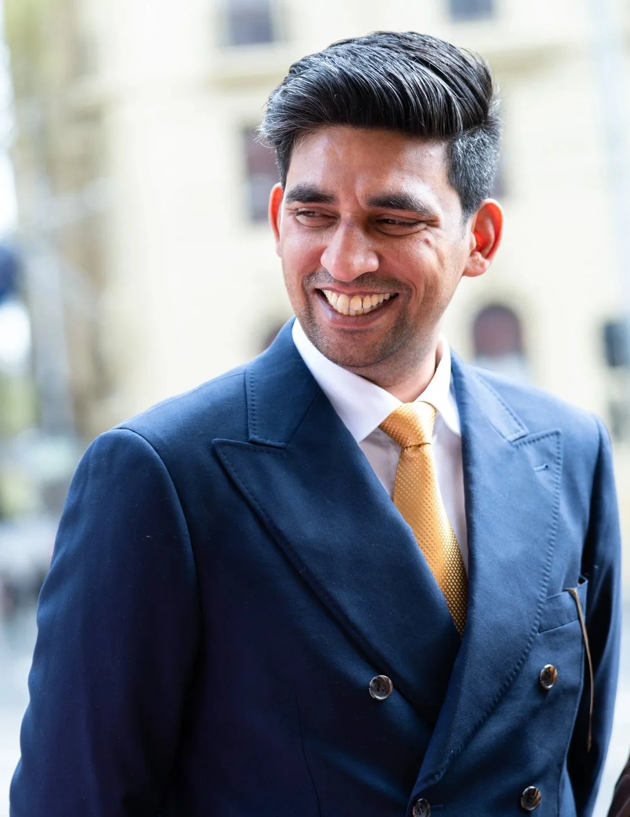 A man with dark, styled hair wearing a navy blue suit, white shirt, and yellow tie, smiling and looking to the side, outdoors in front of a blurred building