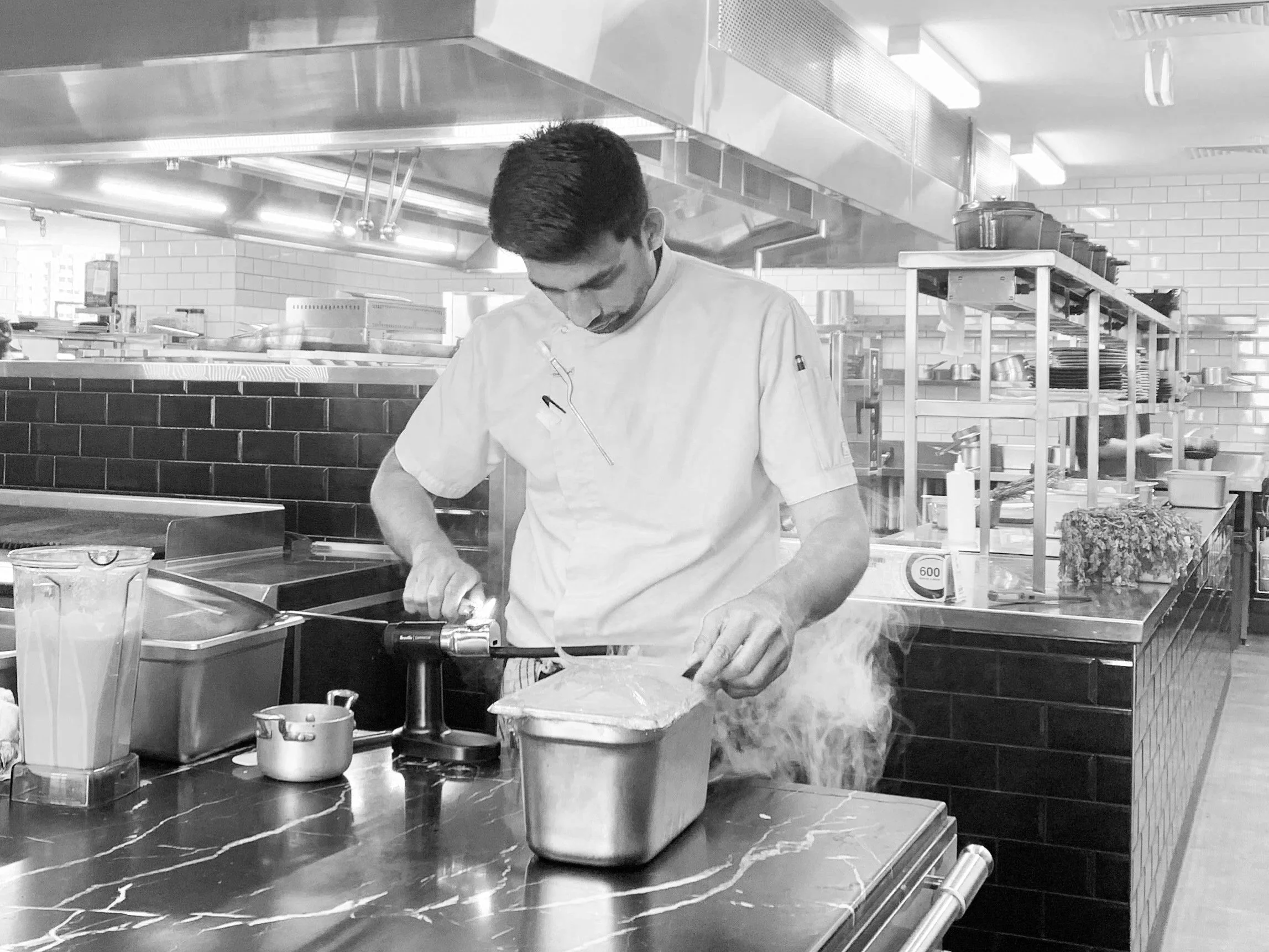 A chef in a commercial kitchen wearing a white chef's coat, working with food or ingredients, surrounded by kitchen equipment, shelves with plates and utensils, in a busy kitchen environment.